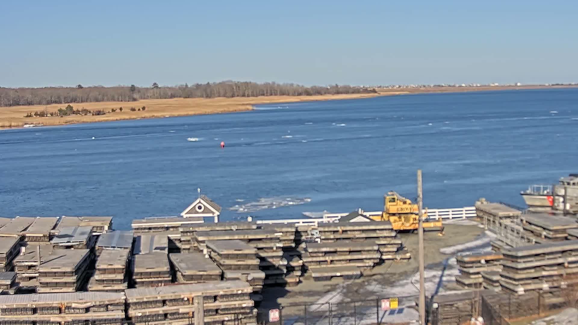 A mostly sunny day shows a body of water with scattered ice floes, a shoreline with stacked rectangular structures, and a yellow crane near a small building.