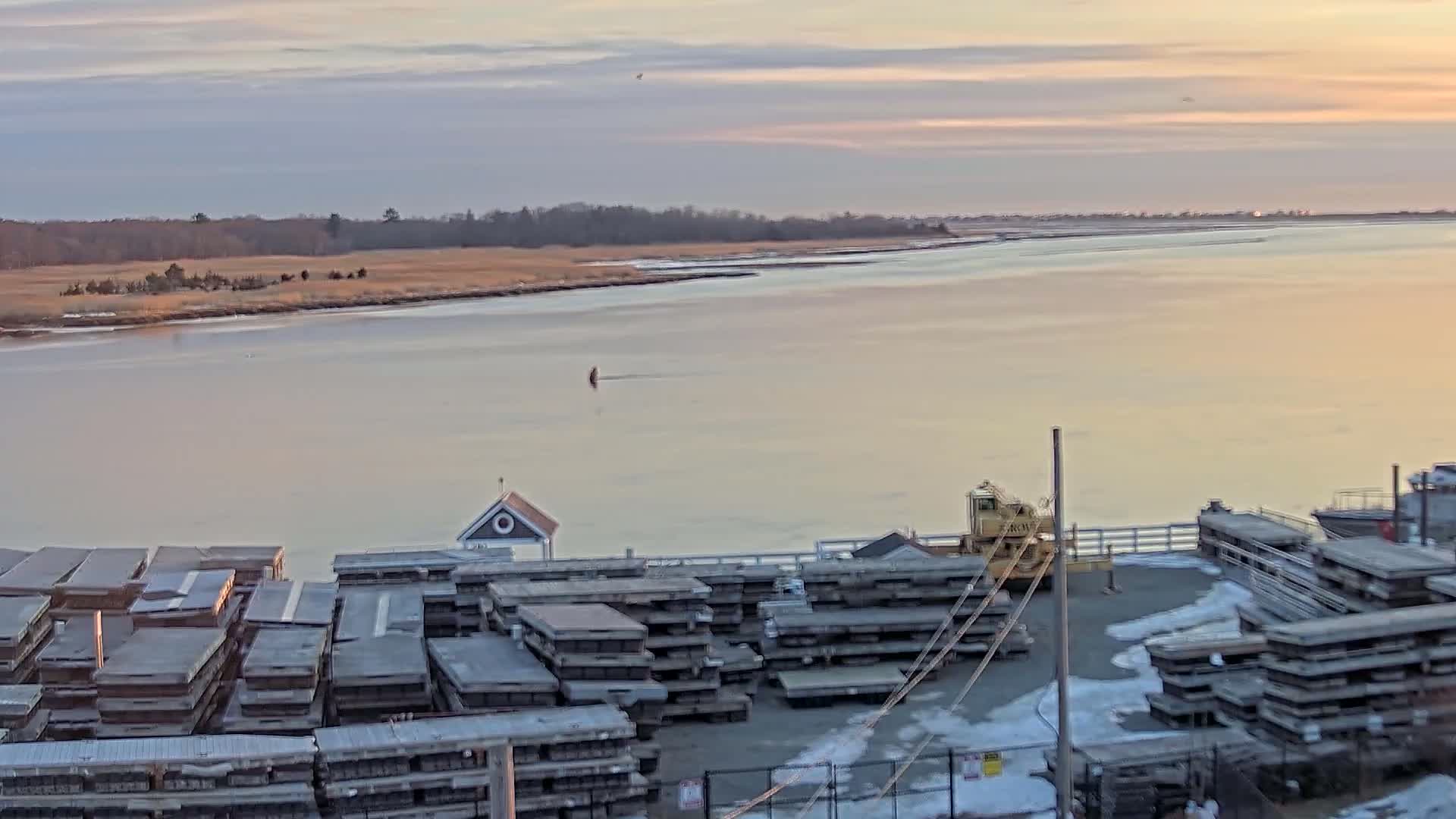 A calm, pale-pink sunset illuminates a wide river with stacks of materials and a small crane on a dock next to a shoreline of muted brown grasses and distant trees.