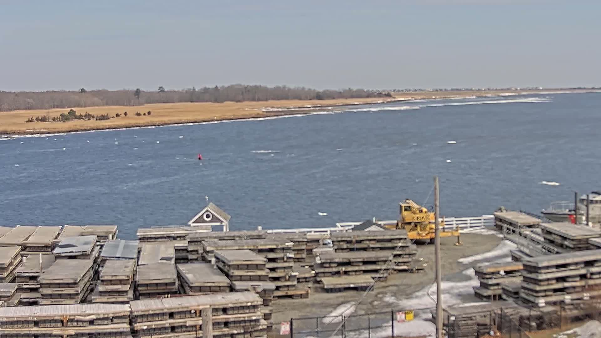 A body of water with a shoreline of brown grasses and trees under a clear sky shows stacks of what appear to be wooden pallets or dock sections near a yellow crane, all adjacent to the water.