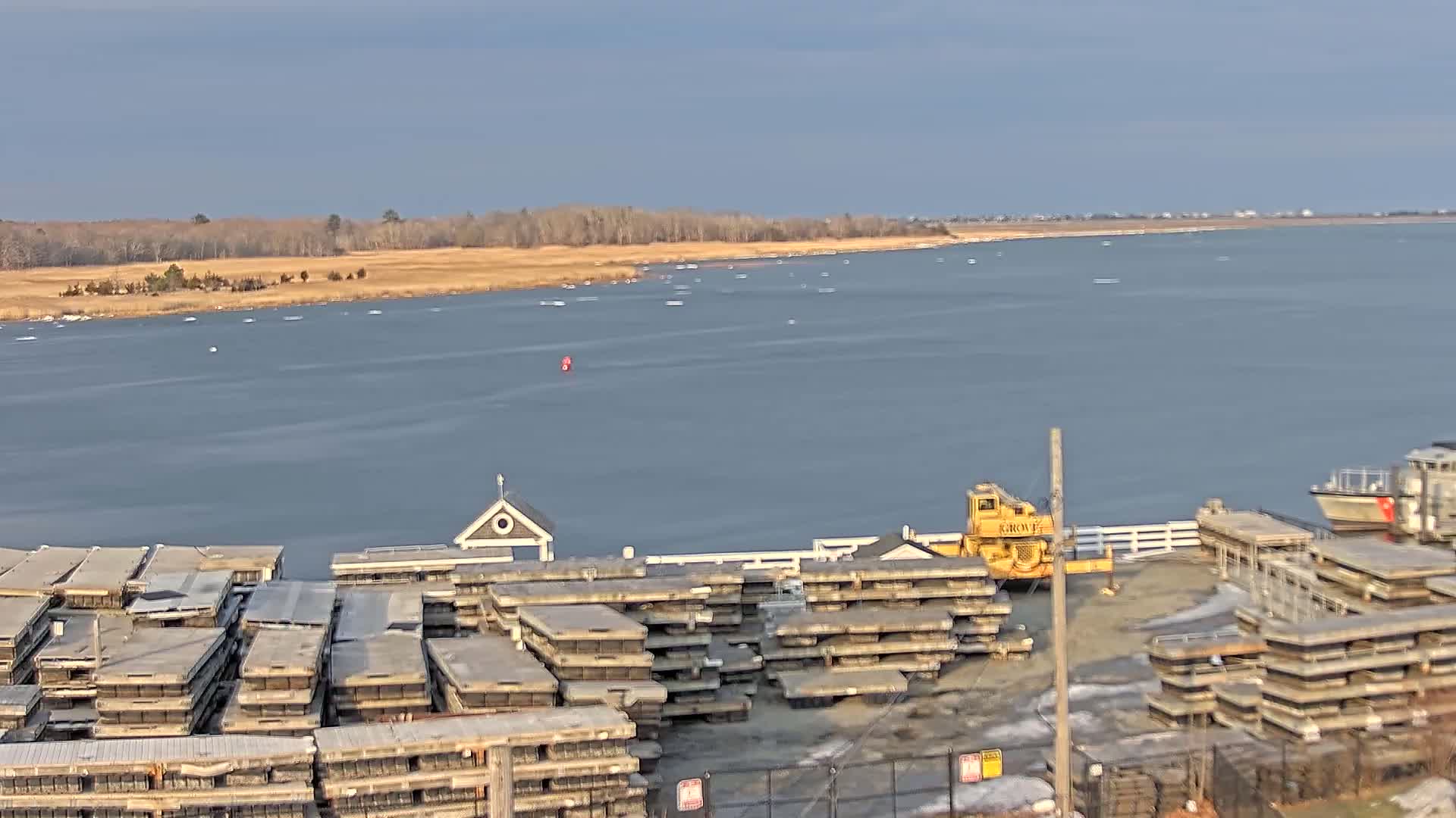 A waterfront area with stacks of what appear to be dock sections, a small building, a yellow crane, a boat, and a calm body of water under a mostly clear sky.