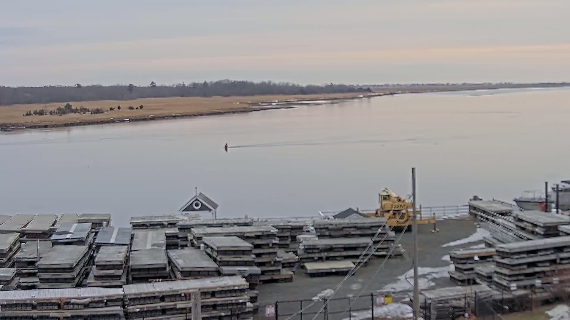 A calm, overcast day reveals a wide river with a small boat in the distance, a yellow crane, and numerous stacked gray floating docks along a shoreline with sparse, brown vegetation.