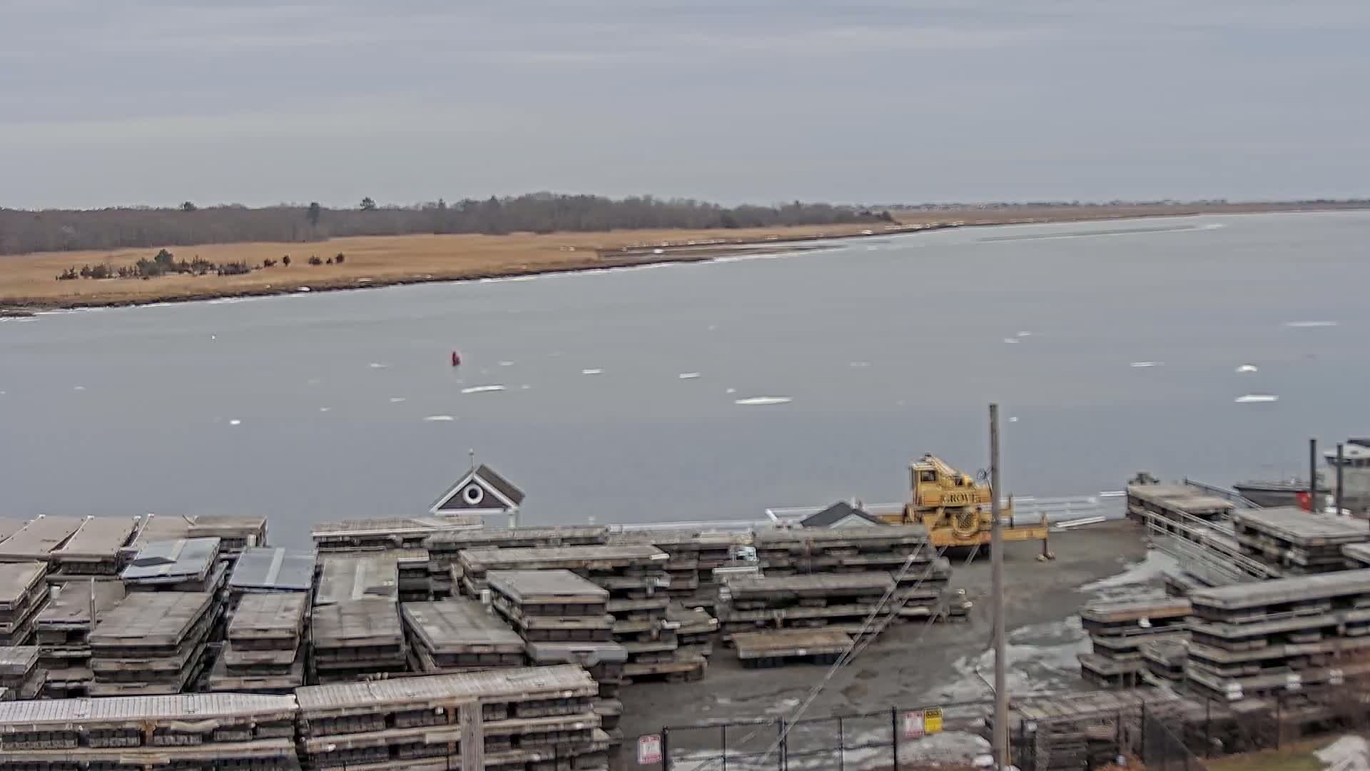 A body of water with scattered ice floes stretches out before a shoreline where stacks of wooden planks are situated next to a yellow crane, under an overcast sky.