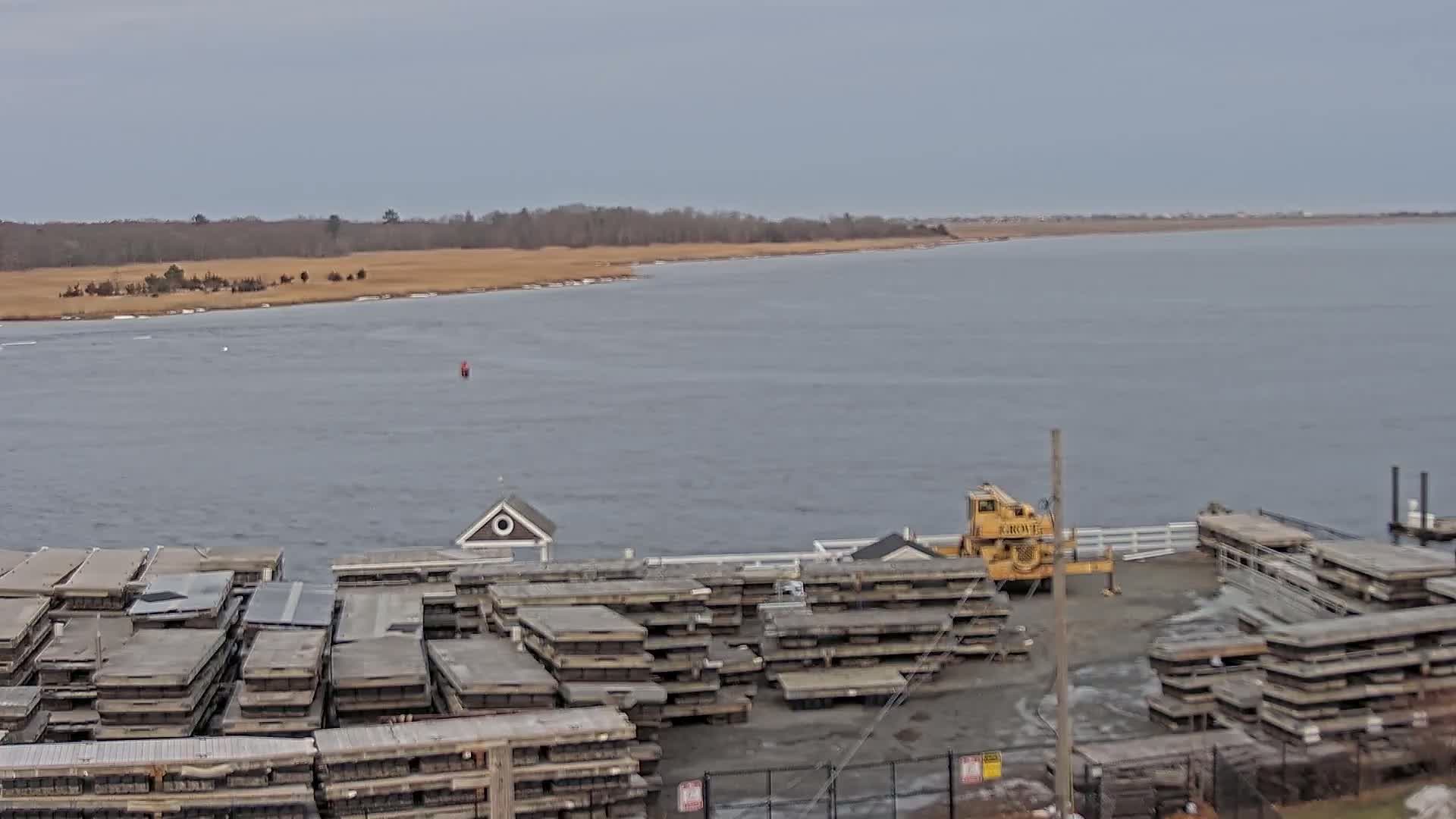 A body of water with a distant shoreline is seen from a high vantage point, showing stacks of what appear to be floating docks near a small structure and a yellow crane under an overcast sky.