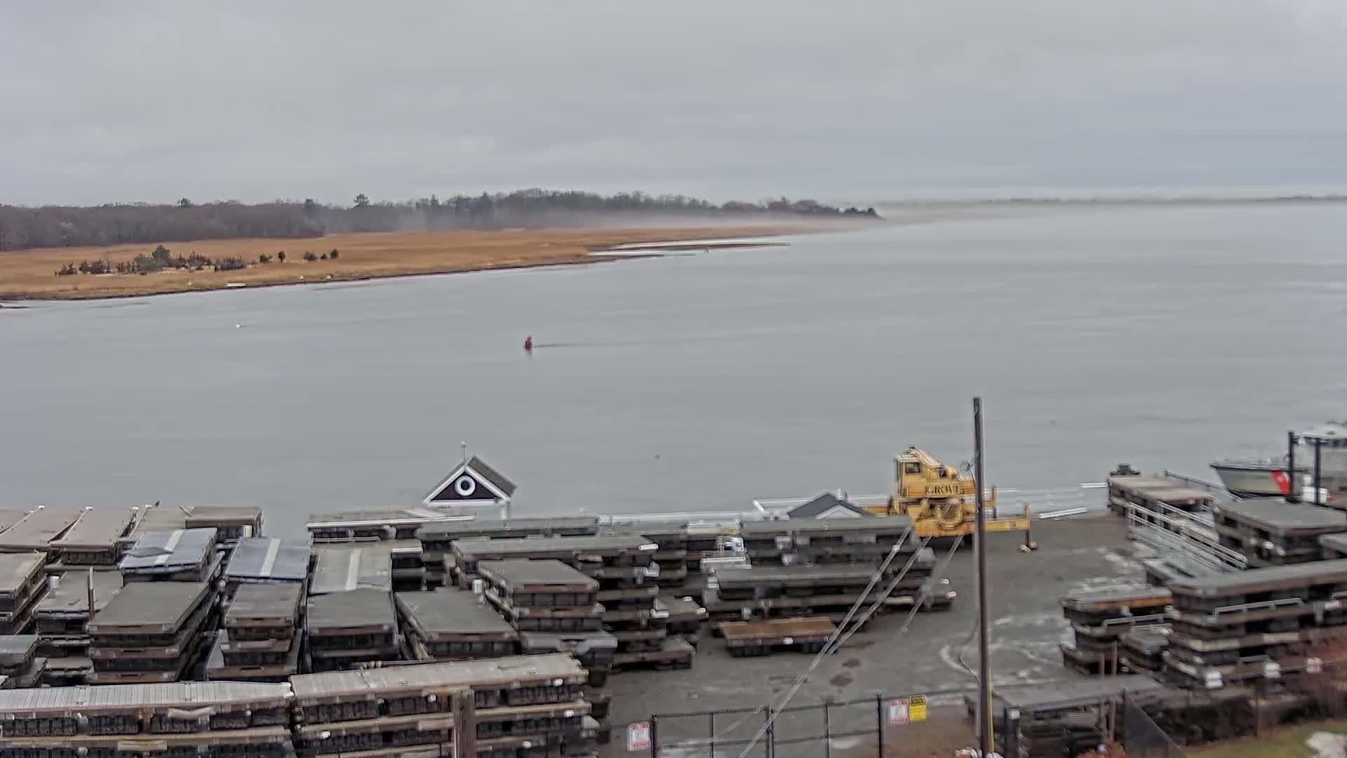 A gray, overcast day shows a body of water with a distant shoreline, a small building, a yellow crane, and numerous stacks of dark gray floating docks.