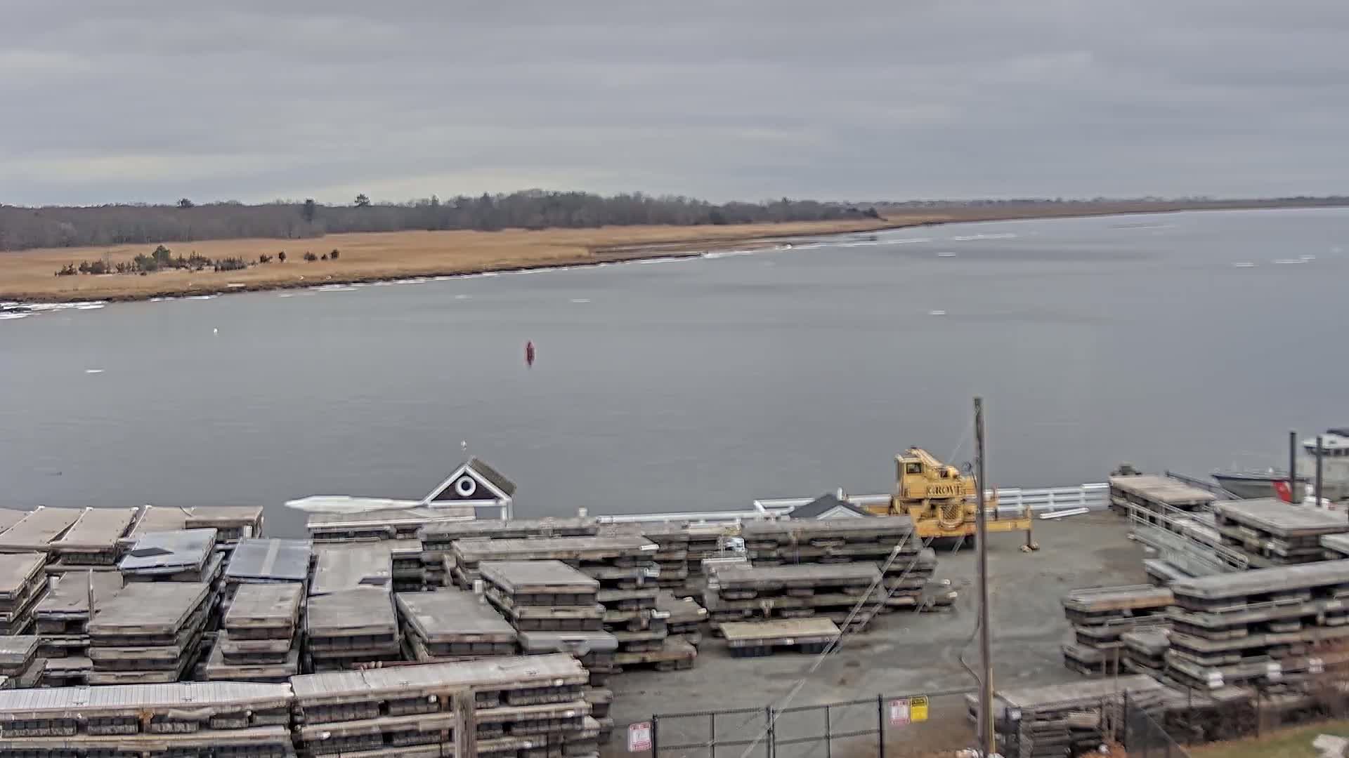A body of water with scattered ice flows under an overcast sky, showing a shoreline with stacked dock sections and a small building next to a yellow crane.