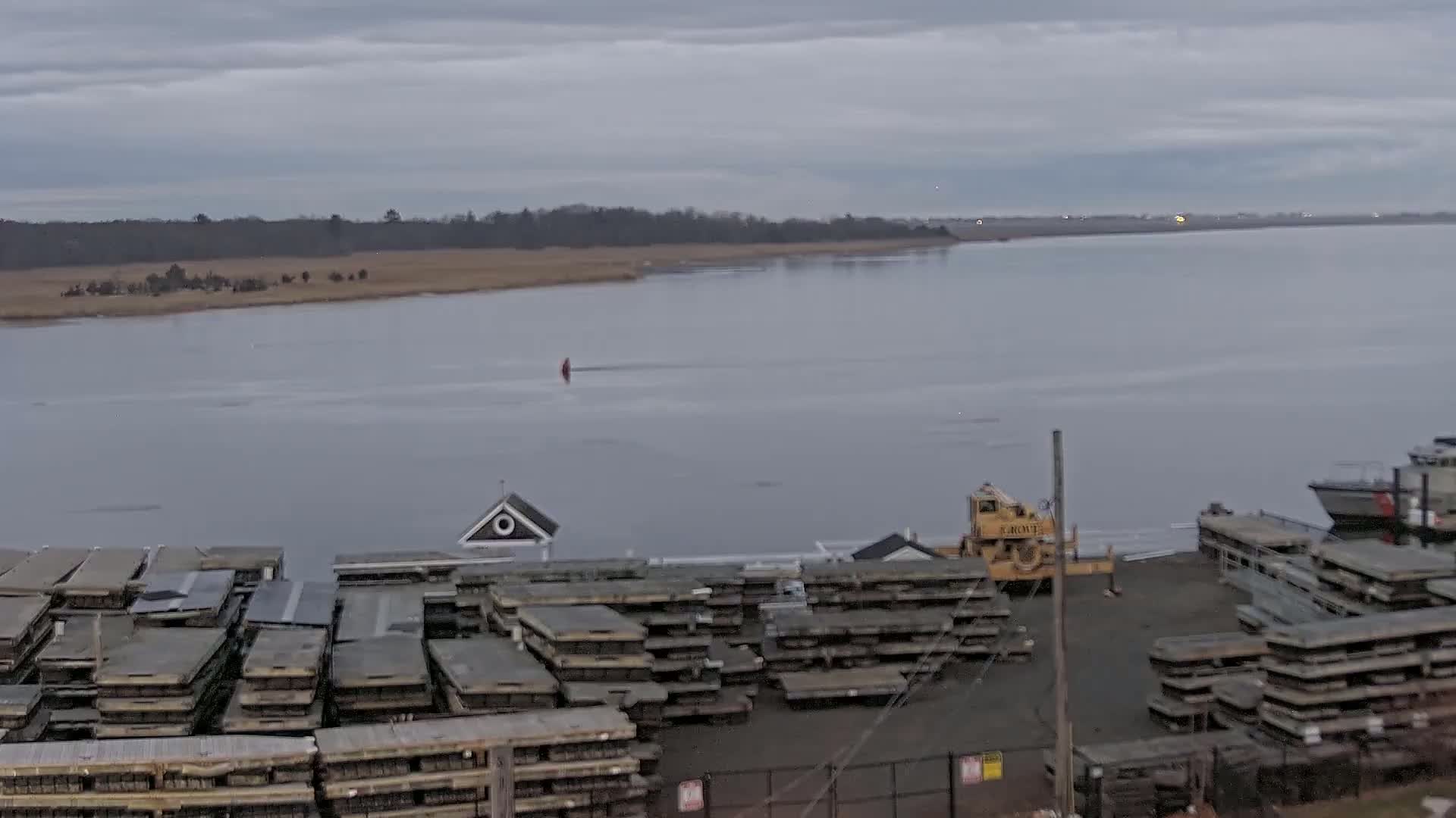 A partly frozen body of water is seen from a high vantage point, showing stacks of floating docks and a small building near a crane and boat on shore under an overcast sky.