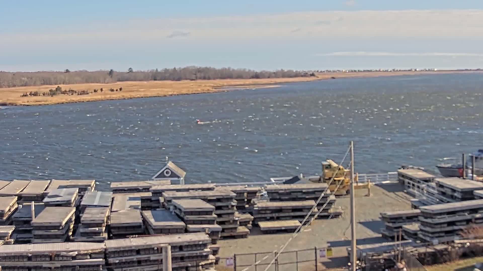A windy day shows a body of water with stacked wooden platforms near a dock, a small building, and a yellow piece of heavy machinery; in the distance are trees and a shoreline.