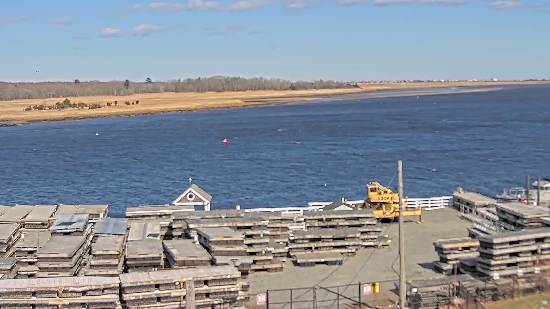 A body of water under a partly cloudy sky is seen from a high vantage point, showing a dock with stacked materials and a small building, along with a yellow crane and a boat.