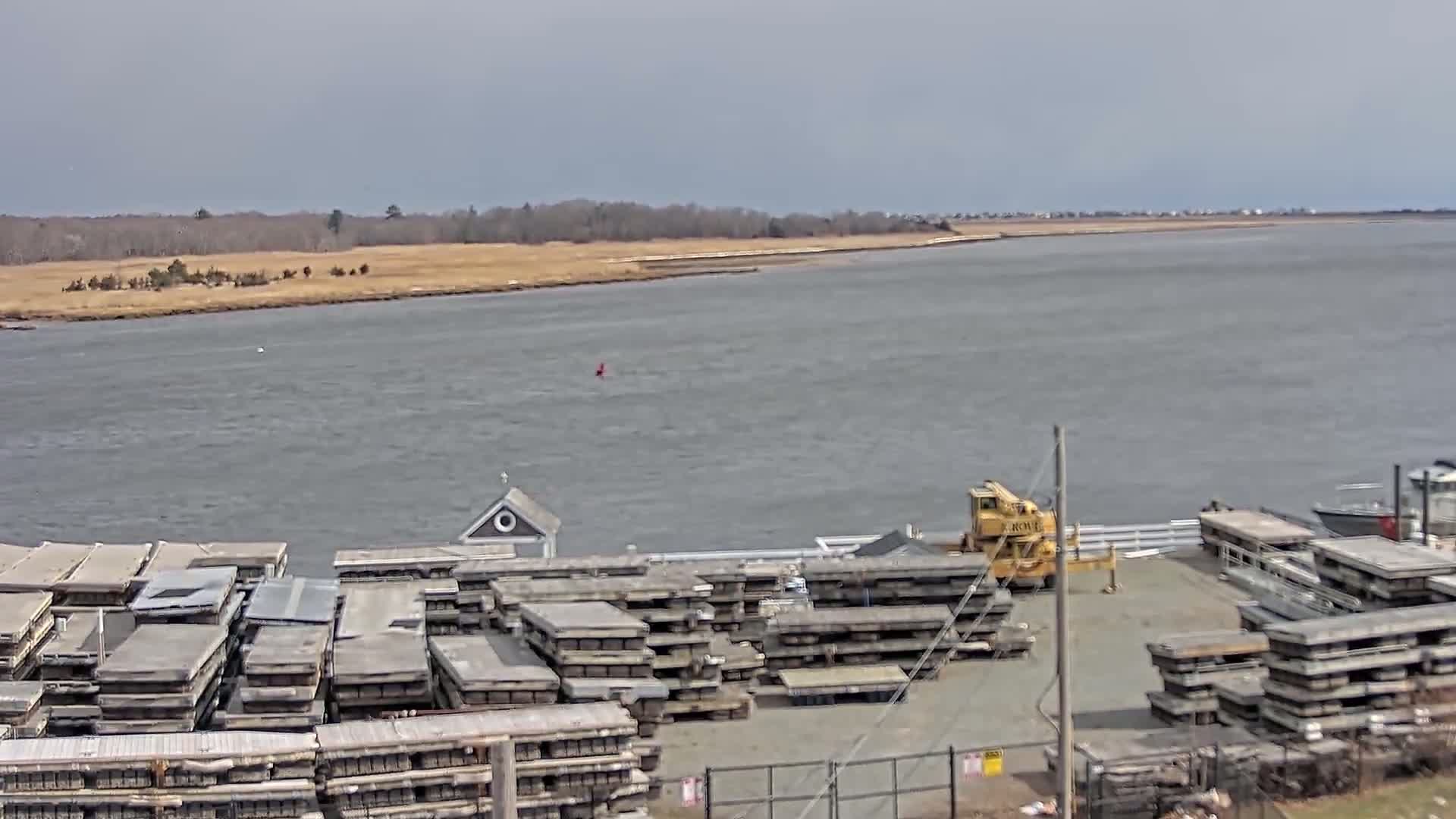 A wide shot of a body of water under an overcast sky shows a dock with stacks of grey rectangular objects and a yellow crane, with a small building and more stacks of the same objects beyond it, all before a marshy shoreline and a line of trees in the distance.