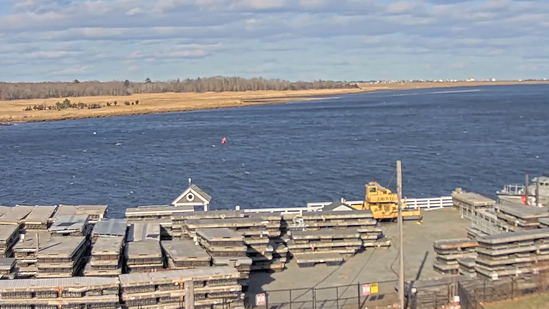 A partly cloudy day shows a body of water with stacks of what appear to be oyster cages and a yellow crane on a dock near a small building, with a marshy area and trees in the background.