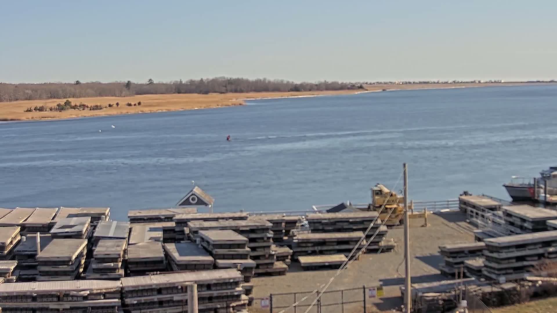 A calm body of water stretches toward a distant shoreline under a clear blue sky, with numerous stacks of what appear to be floating docks located in the foreground.