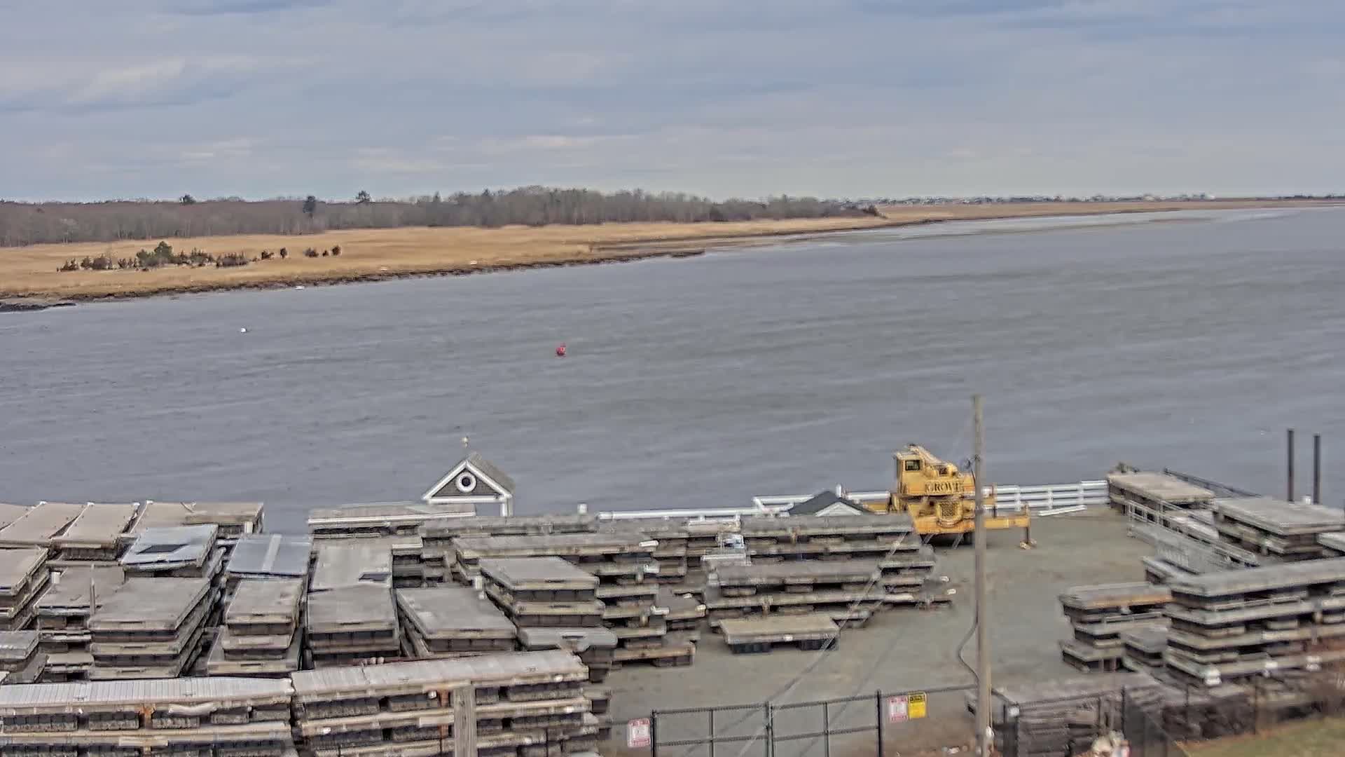A wide shot shows a body of water under a cloudy sky, with numerous stacked flat pieces of material near a yellow crane on a dock next to a marshy shoreline.