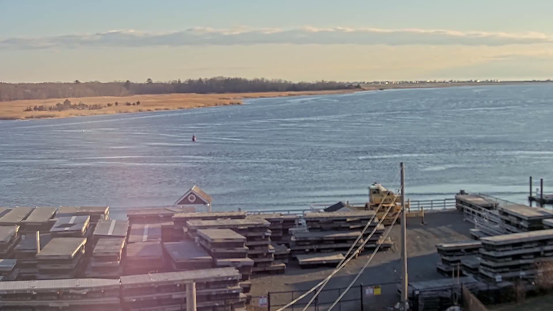 A body of water with light chop under a mostly clear sky shows a shoreline with stacked wooden planks and a yellow piece of machinery near a small building.