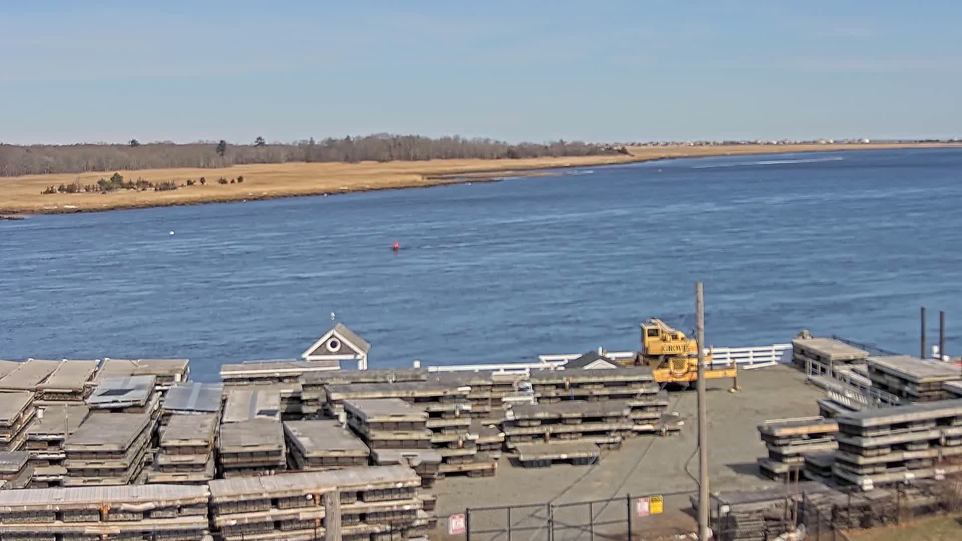 A body of water under a clear blue sky stretches beyond a dock where numerous stacked platforms are stored, next to a yellow crane.