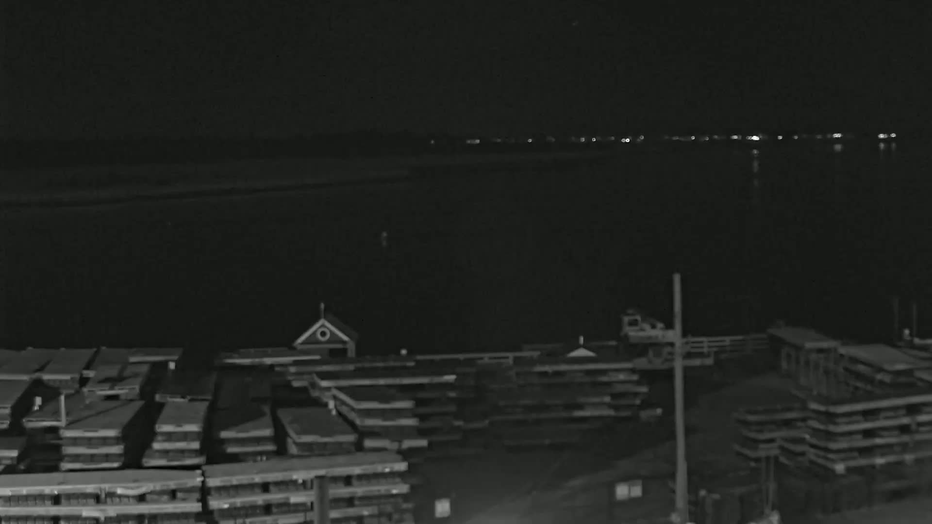 A nighttime, low-light view shows a dark body of water with faintly visible lights across it, and a dock with numerous stacked structures in the foreground.