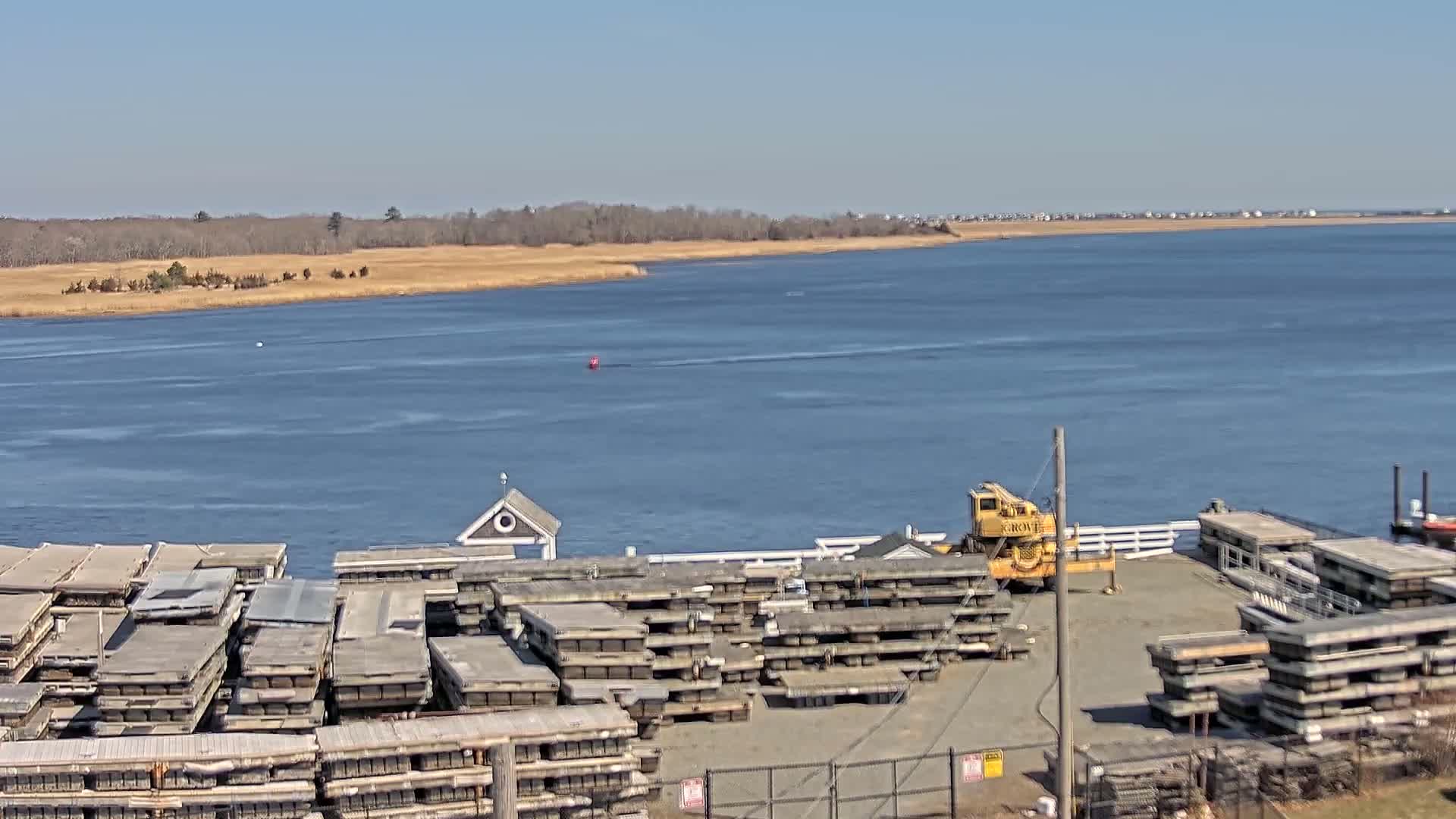 A body of water stretches to a distant shoreline under a clear blue sky, with numerous stacked floating docks and a crane visible on the foreground shore.