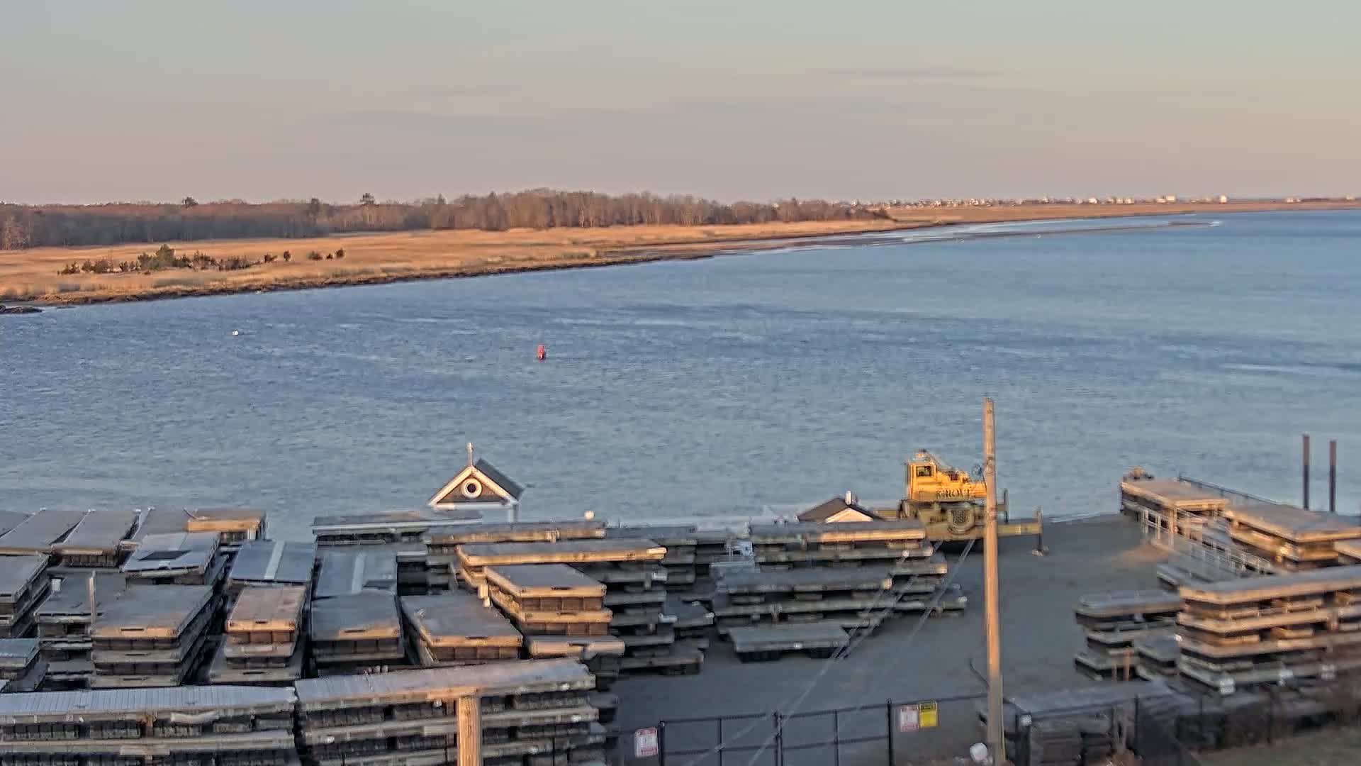 A calm body of water stretches out to a distant shoreline of trees and buildings under a clear, pale sky, with a dock in the foreground containing numerous stacked floating platforms and a small yellow crane.