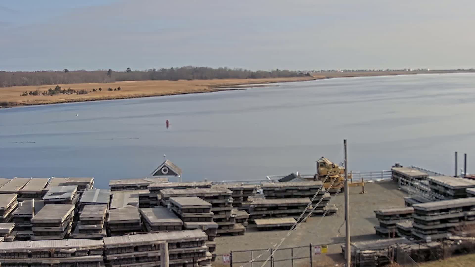 A calm body of water is seen beyond a dock where numerous stacked concrete slabs are stored, a small building, and a piece of heavy equipment.