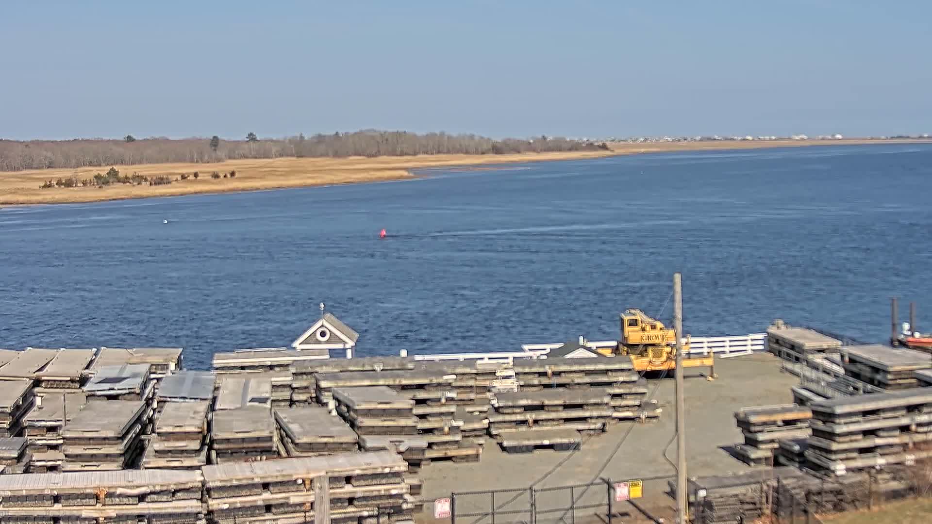 A body of water stretches to a distant shoreline under a clear blue sky, with numerous stacked floating docks and a crane at the foreground.