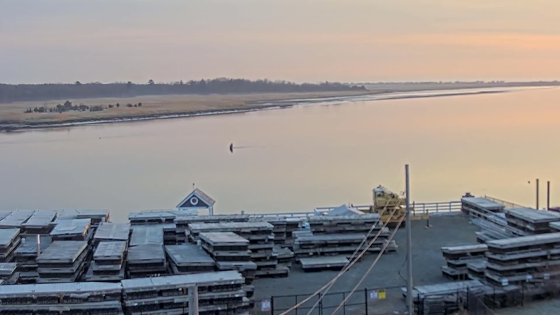 A calm, pale-pink sunrise illuminates a wide river, with stacks of gray rectangular objects and a yellow crane visible on a dock in the foreground.