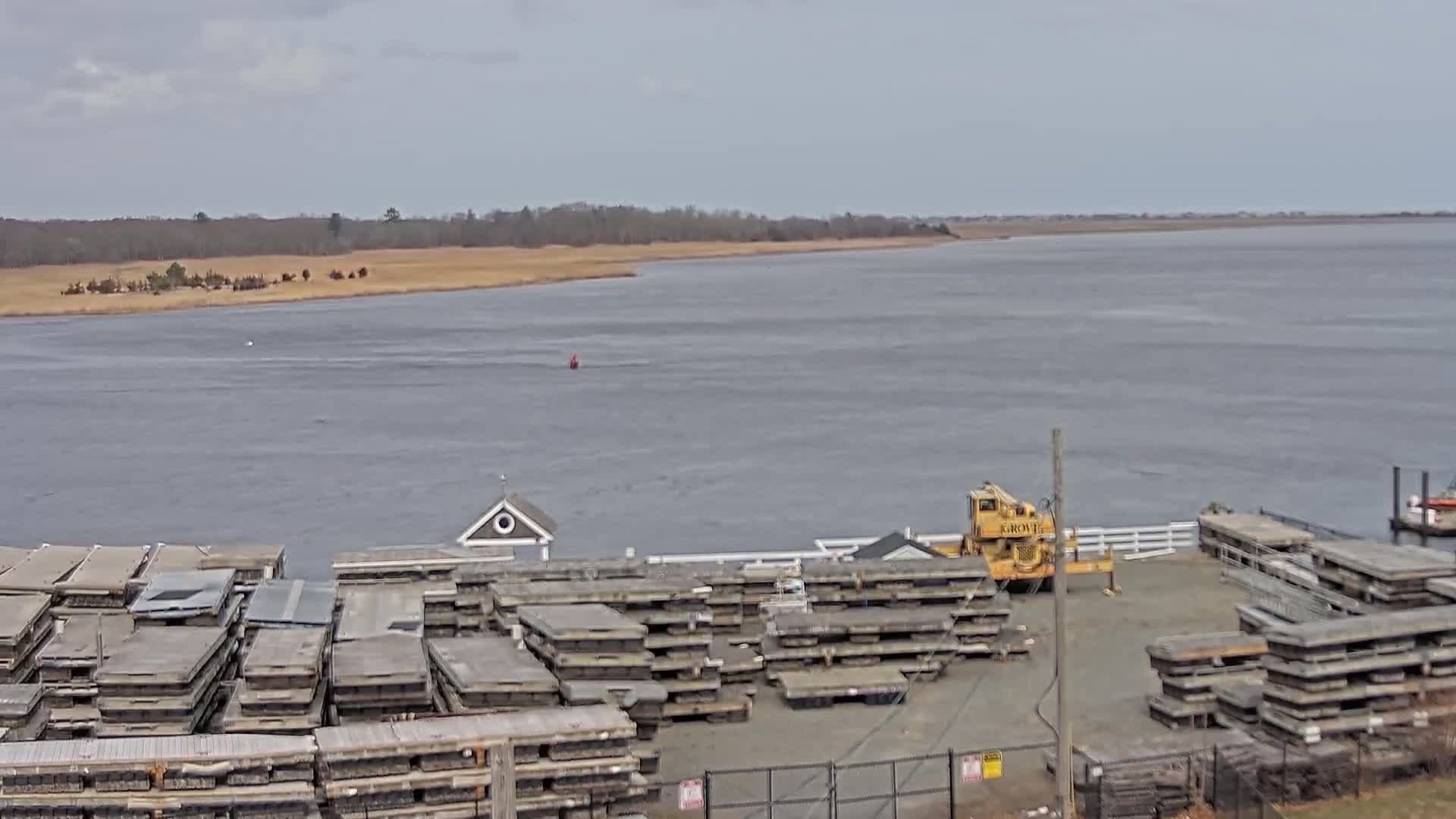 A wide, gray body of water stretches behind a yard filled with stacked, gray wooden planks and a yellow crane, all under an overcast sky.
