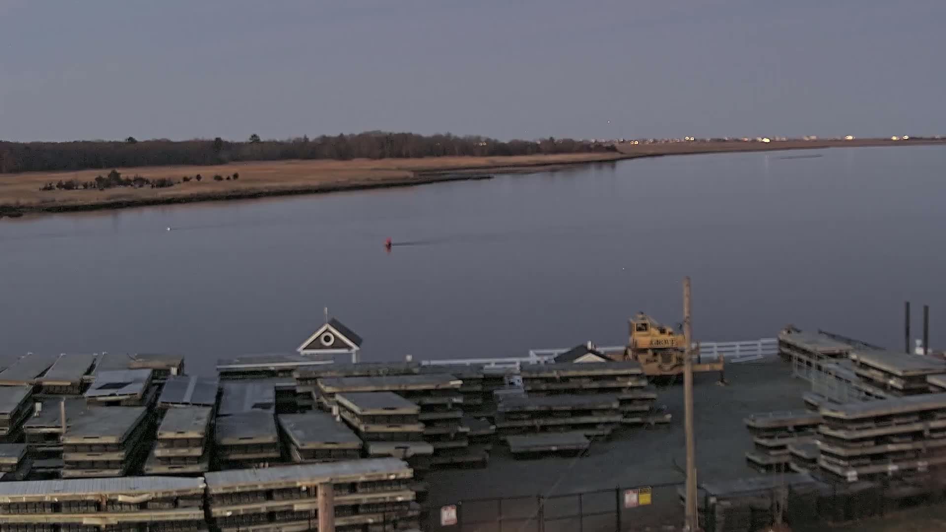 A calm, dusky-lit river is seen from an elevated vantage point, showing a dock with stacked materials and a small building, with a distant shoreline and a bit of land in the background.