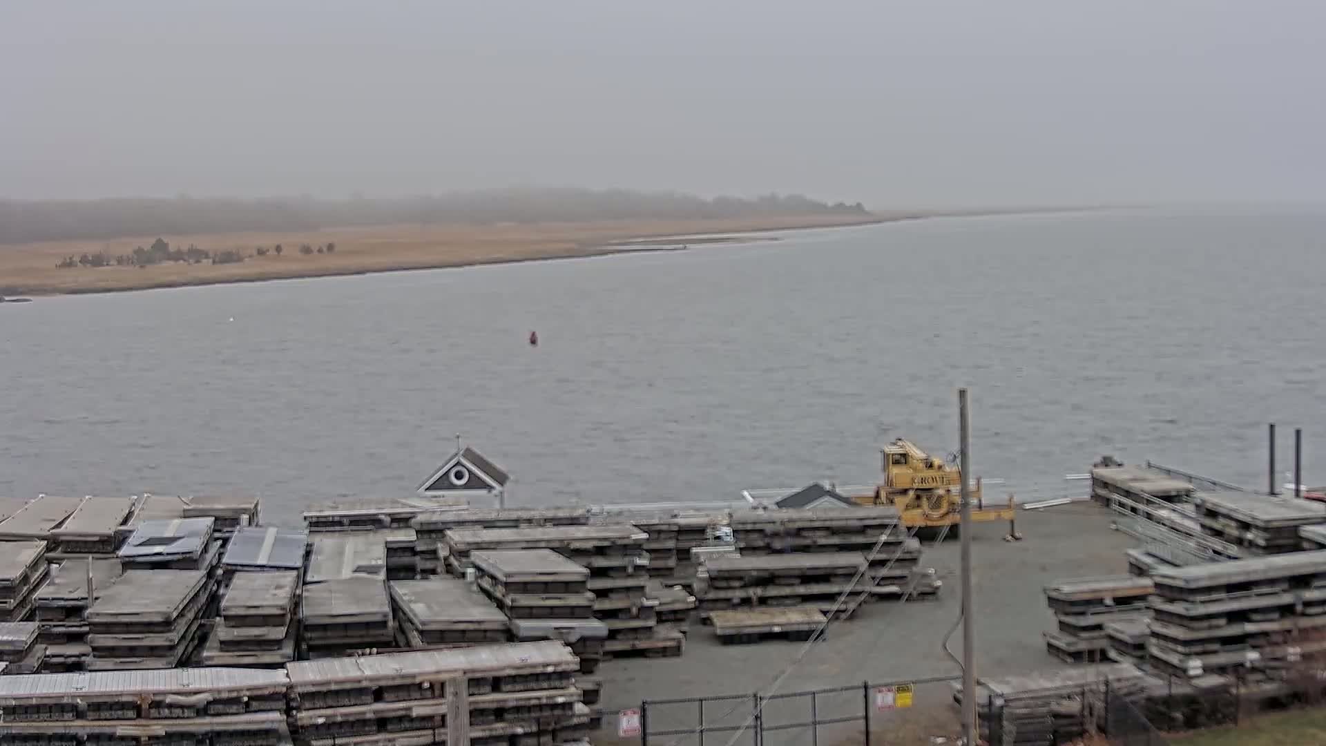 A body of water under an overcast sky is seen from a high vantage point, showing a shoreline with stacked, grey floating docks and a yellow crane.