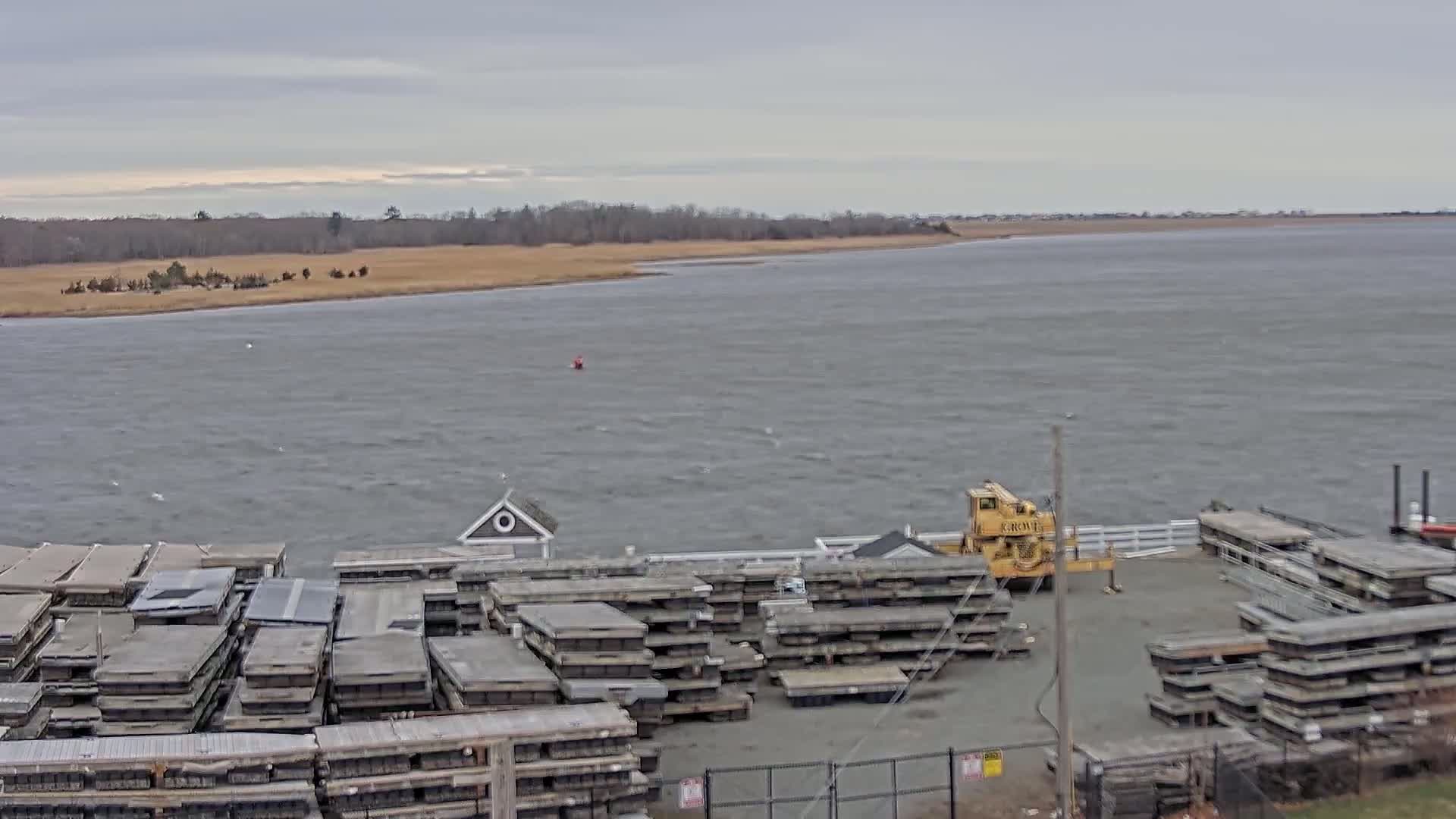 A choppy body of water stretches toward a distant shoreline under a cloudy sky, with numerous stacked, gray platforms and a yellow crane visible in the foreground.