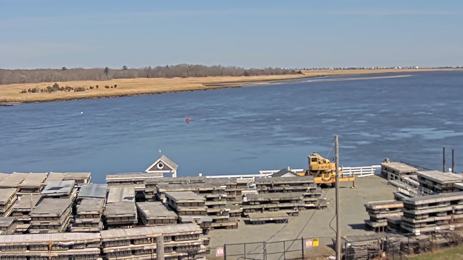 A wide body of water under a clear blue sky is seen beyond a dock holding stacks of what appears to be wooden planks and a yellow crane.