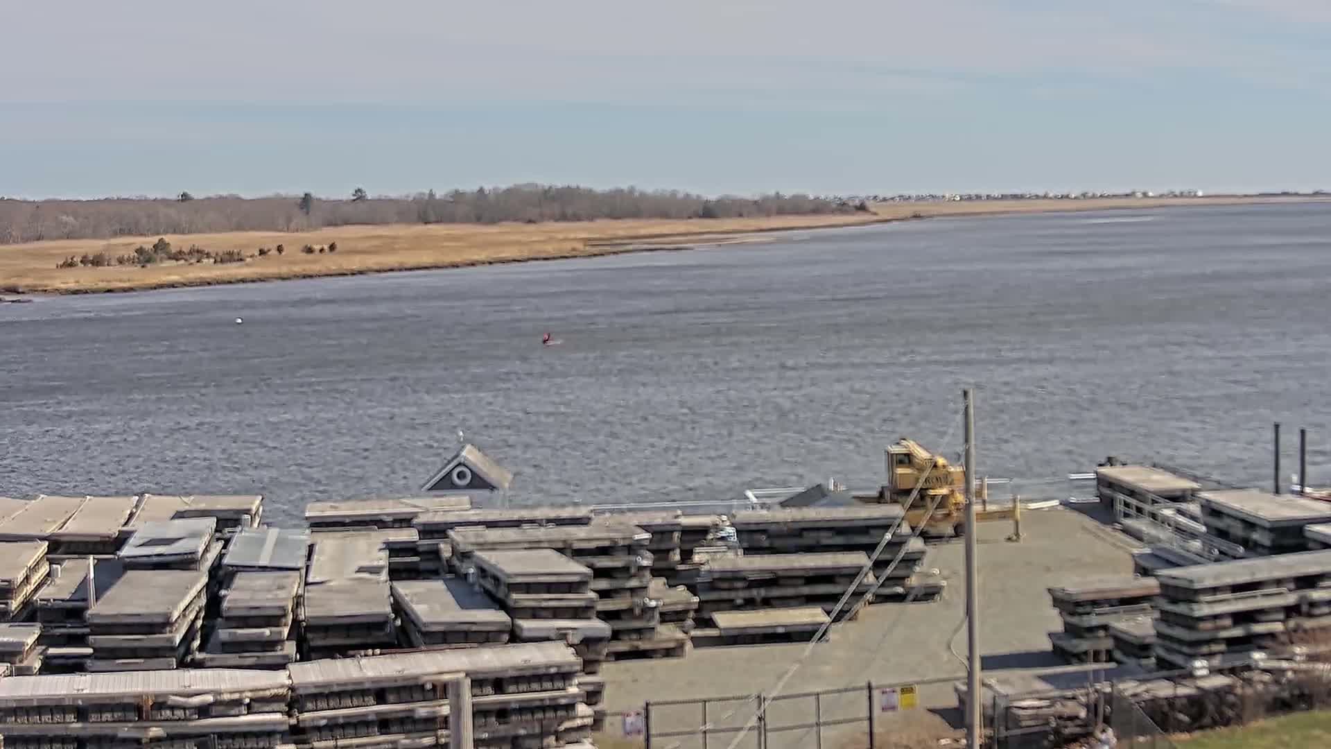 A body of water with a shoreline of brown grasses and trees is seen under a mostly clear sky, with stacks of gray rectangular objects and a crane near the water's edge.