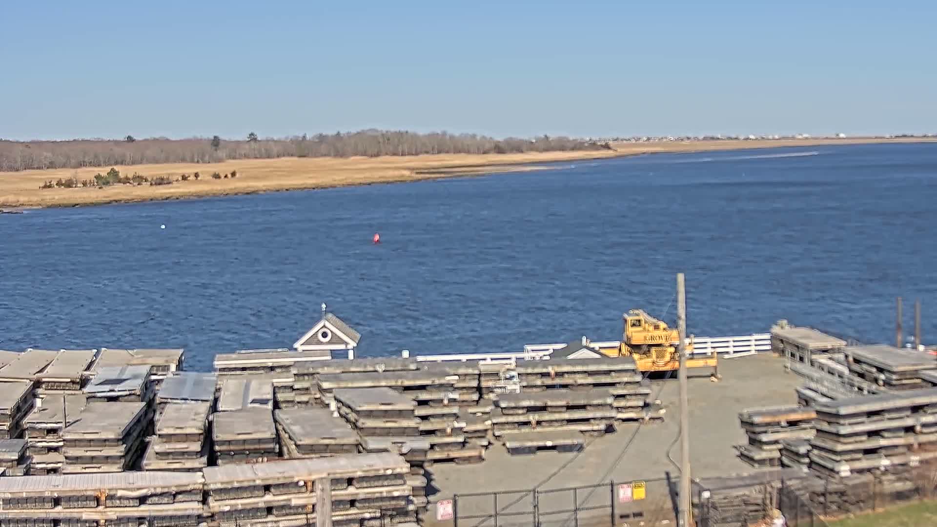 A yellow crane sits near stacks of gray rectangular objects by a body of water on a clear, sunny day, with a treeline visible in the distance.