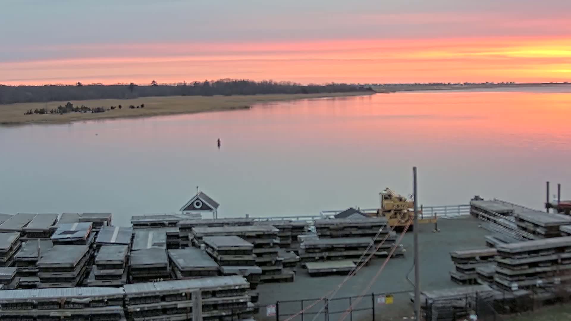 A calm, pink-hued sunrise over a tranquil body of water, reflecting the sky, with numerous stacked floating docks and a small crane visible in the foreground.