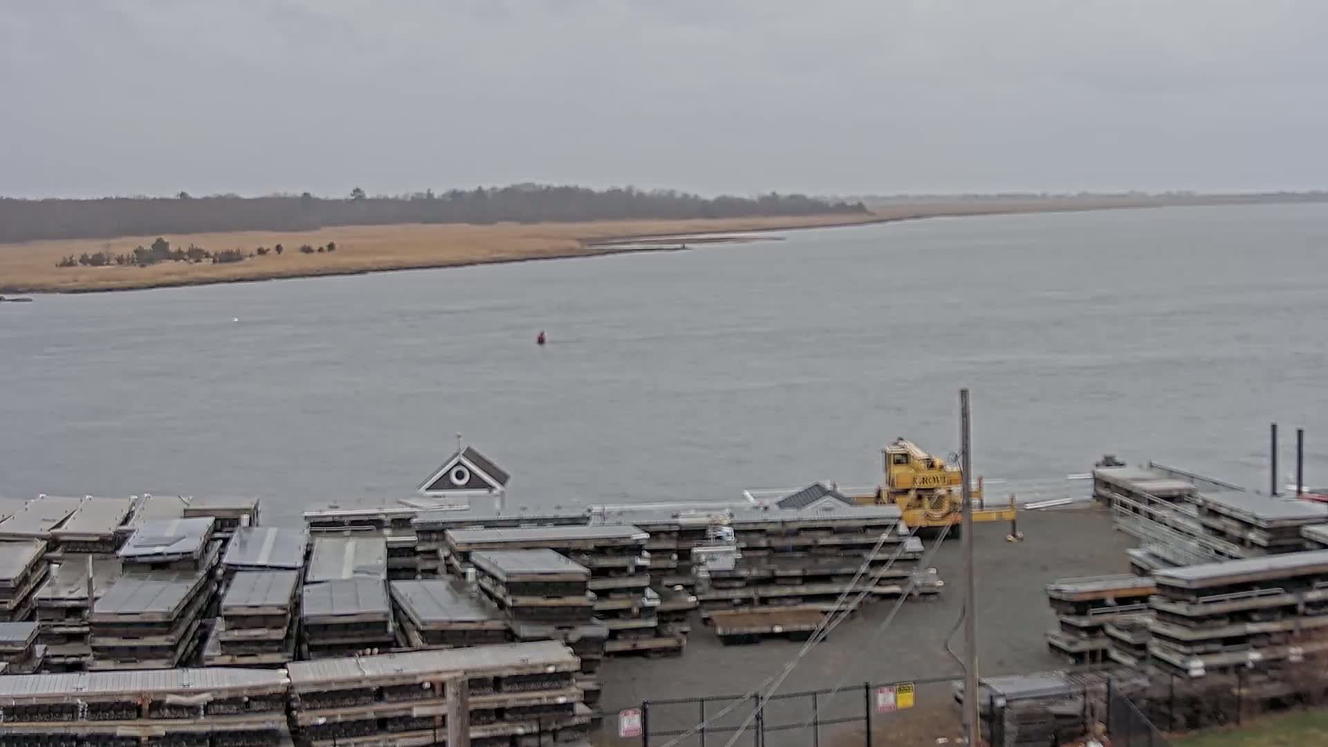 A wide shot of a harbor on an overcast day shows a body of water, stacks of what appear to be floating docks, and a yellow crane.