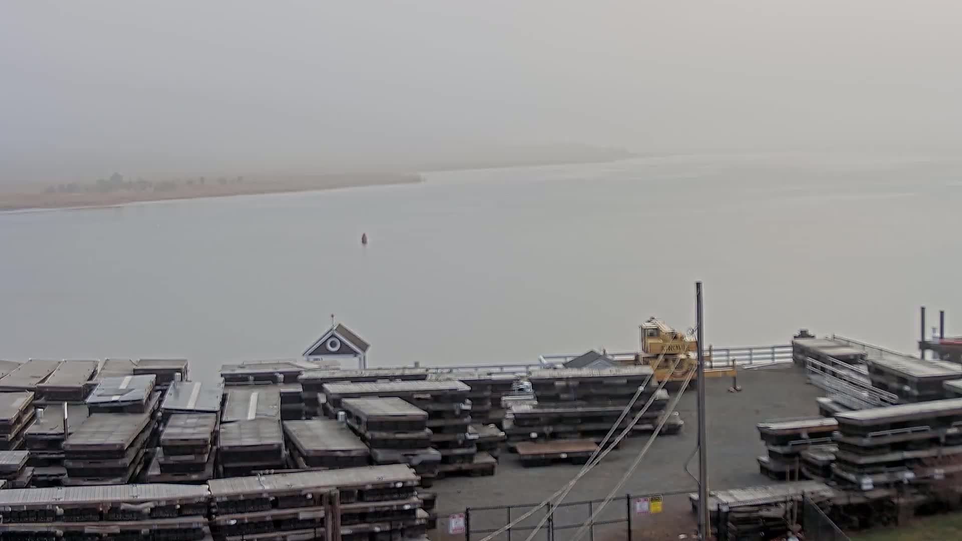 A foggy waterfront scene shows a body of water with stacked, dark-colored floating docks, a small building, a yellow crane, and a distant shoreline.