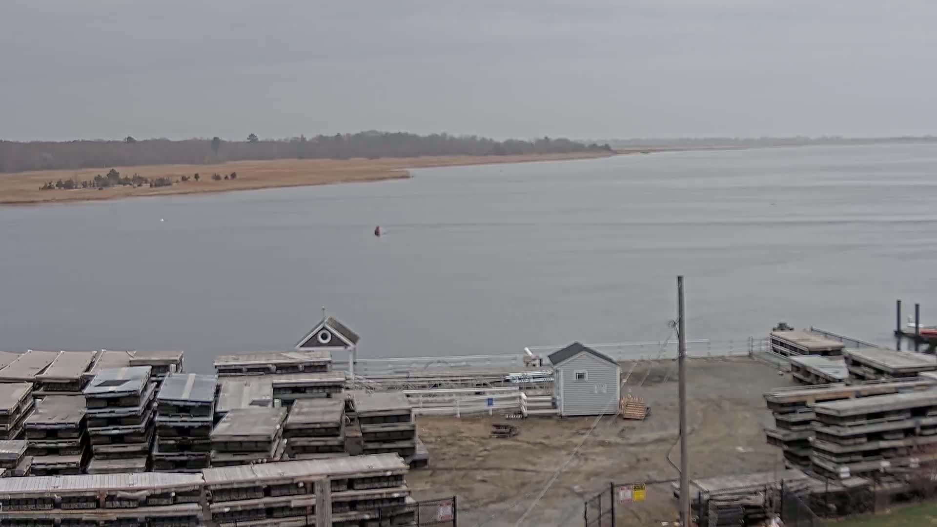 A wide shot of a calm, gray body of water under an overcast sky shows a dock with stacked floating docks and two small buildings, one with a cupola, along a shoreline with sparse, low-lying vegetation.
