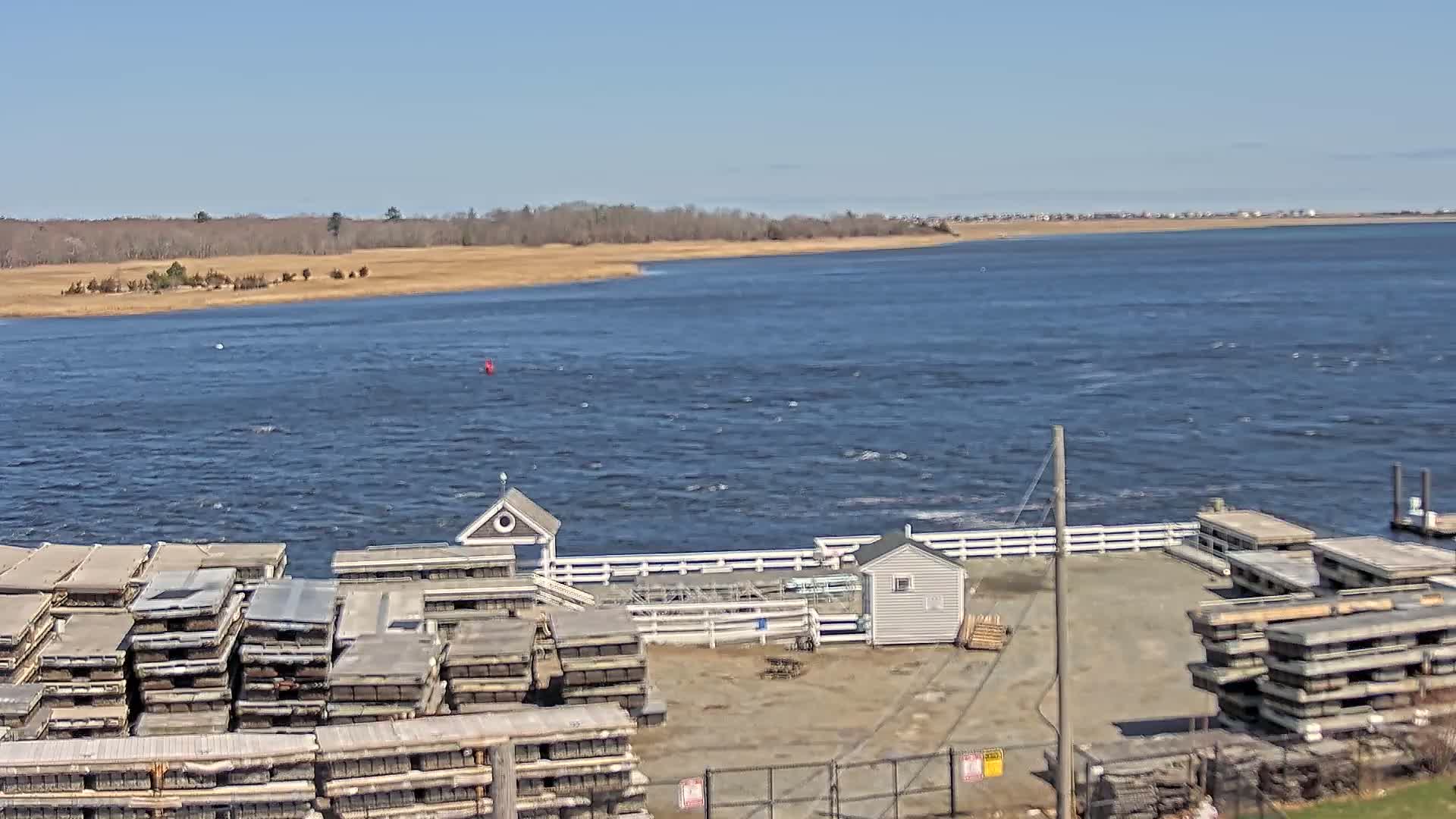 A dock with stacked floating platforms and small buildings sits beside a choppy body of water under a clear blue sky.