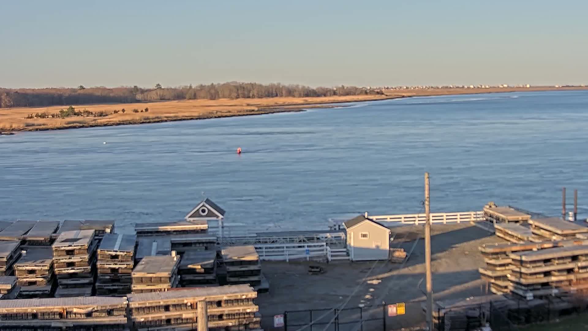 A calm body of water stretches out to a distant shoreline under a clear, bright sky, with a small dock containing several stacked structures in the foreground.