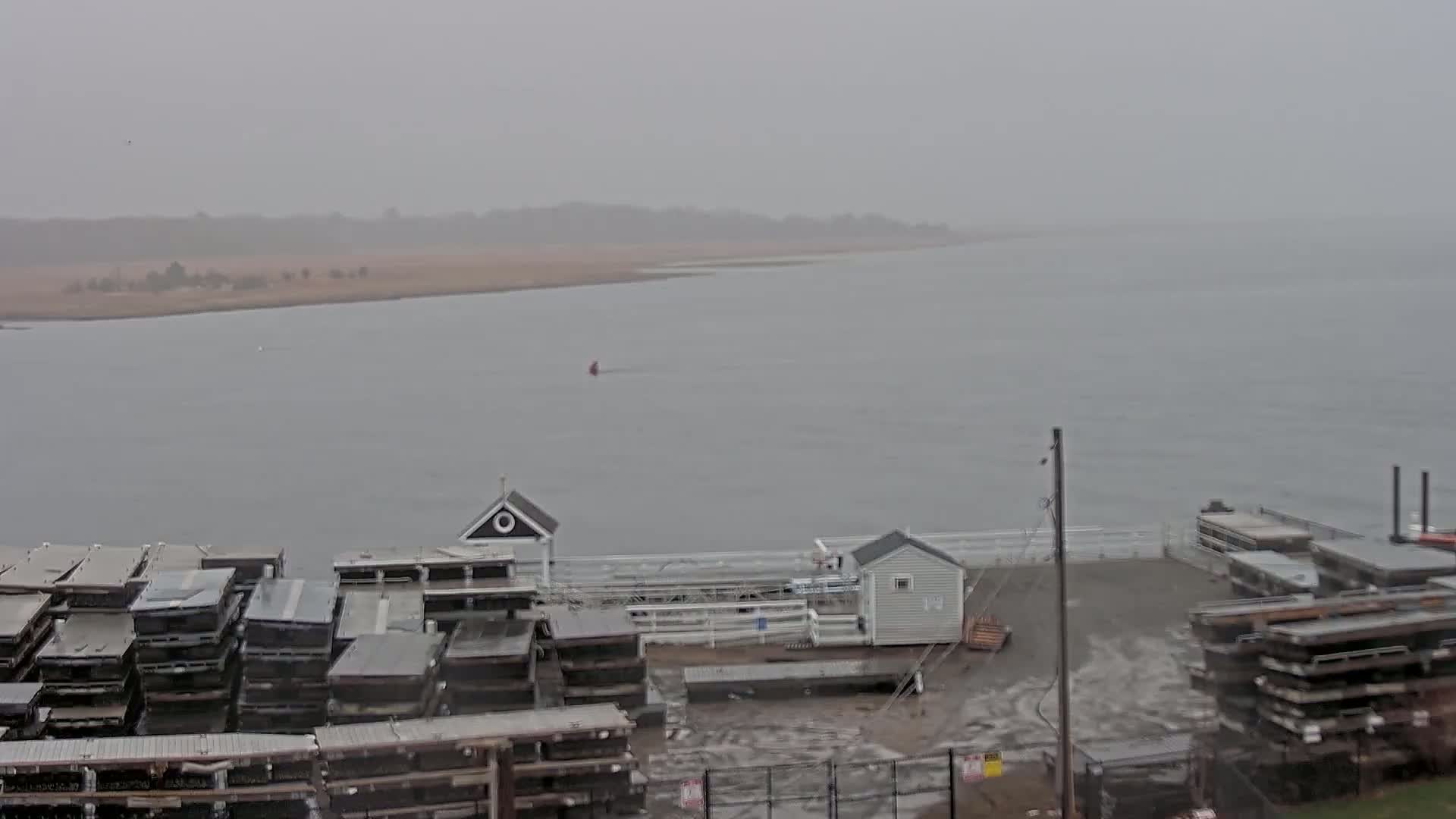 A hazy, overcast day reveals a waterfront scene with stacked boat hulls and small buildings along a shoreline, overlooking a calm body of water extending to a distant, flat landmass.