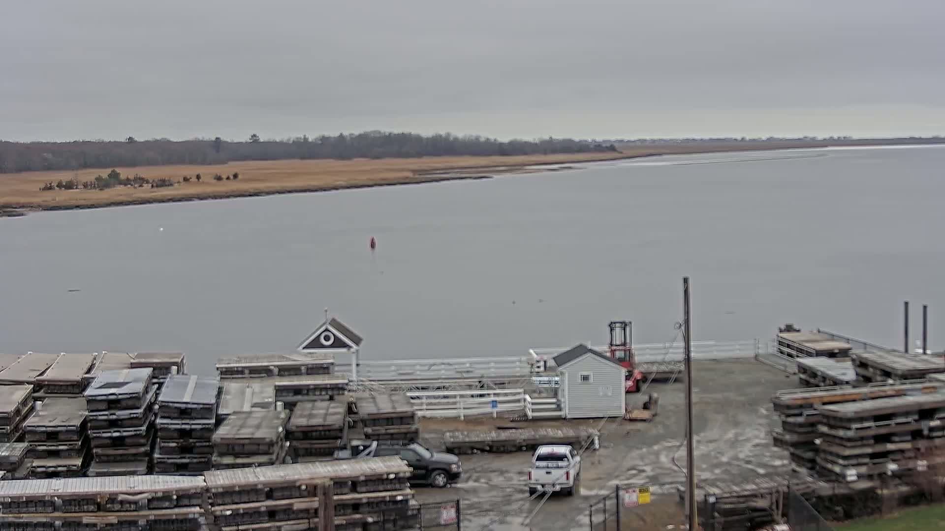 A calm, gray day shows a waterfront scene with stacks of what appear to be boat docks, two small buildings, several vehicles, and a body of water with a flat, marshy shoreline in the background.