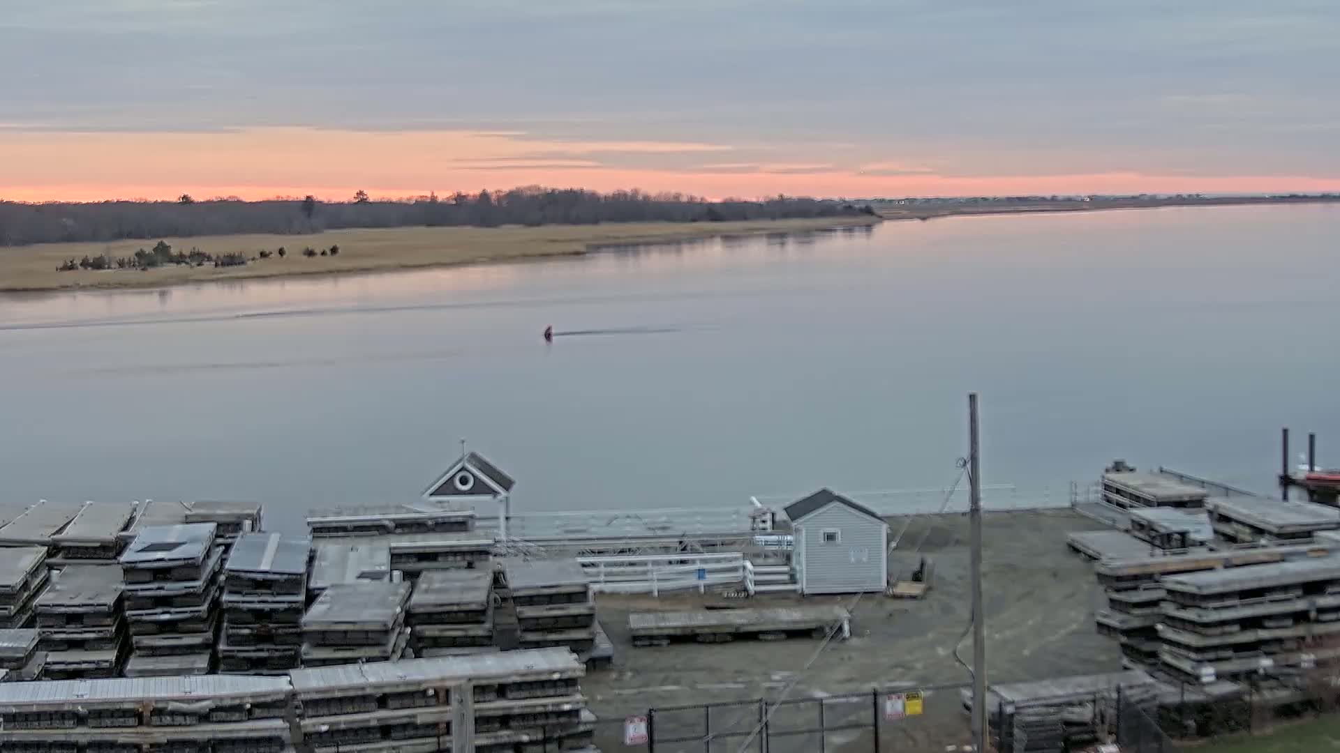 A calm body of water at sunset is seen from an elevated vantage point, showing stacked docks, small buildings, and a single small vessel in the distance.