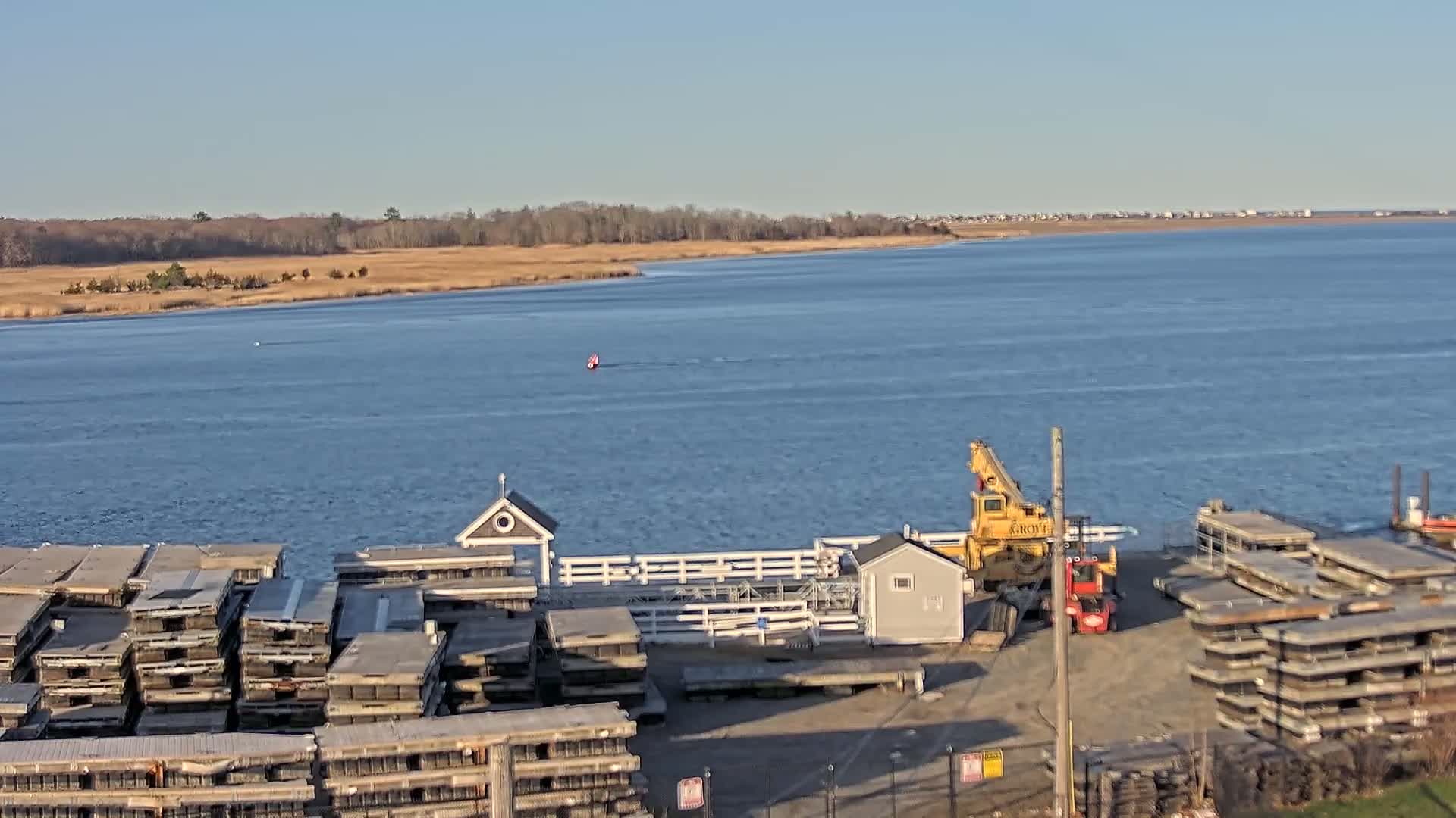 A body of water under a clear, sunny sky shows a small boat in the distance, a crane and a forklift near a dock with stacked floating platforms, and a shoreline with brown marshland and trees in the background.