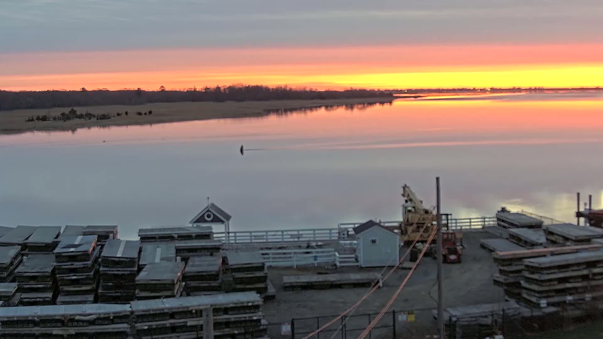 A calm body of water reflects a vibrant sunrise over a treeline, with a dock containing stacked materials and a crane visible in the foreground.
