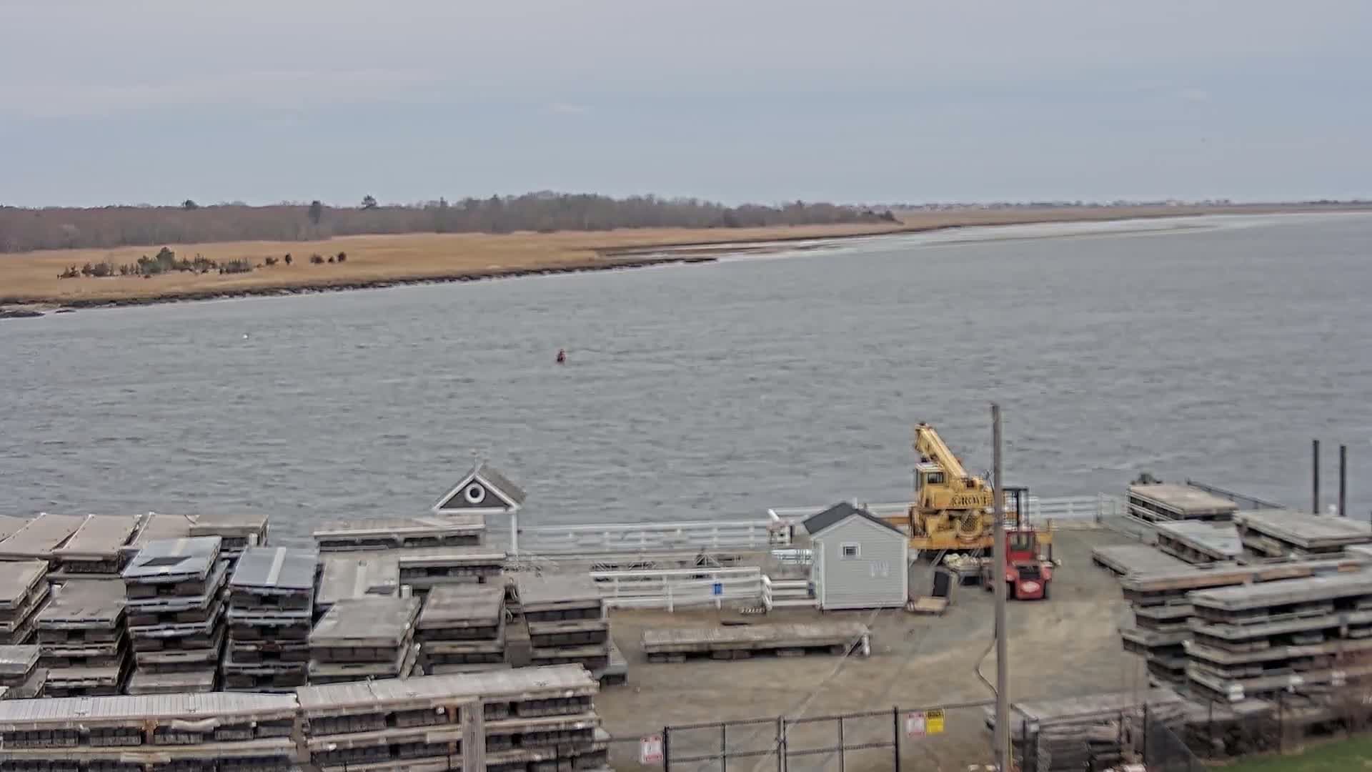 A body of choppy water is seen from a high vantage point, with a dock in the foreground containing stacked flat pieces of wood, a small building, a crane and forklift.