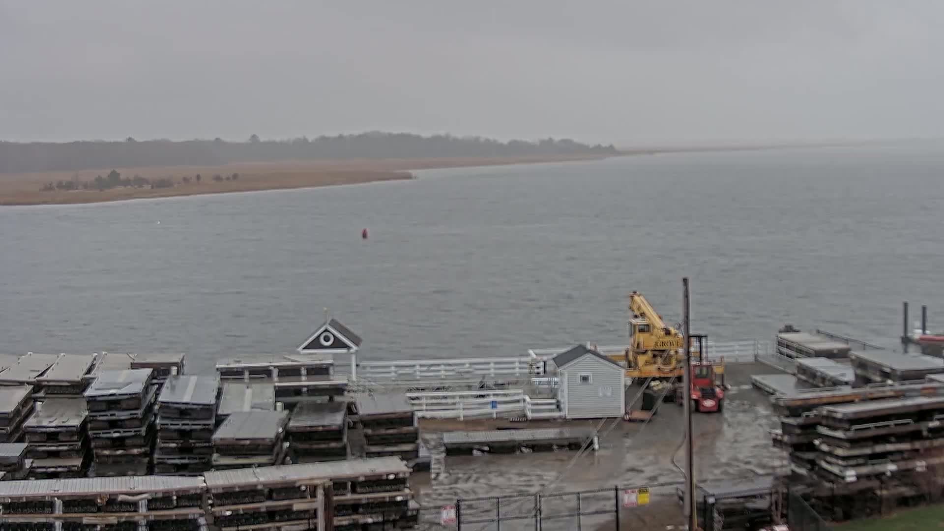 A gray, overcast day shows a body of water with a shoreline in the distance; in the foreground are a dock, a crane, and stacks of dark gray materials.