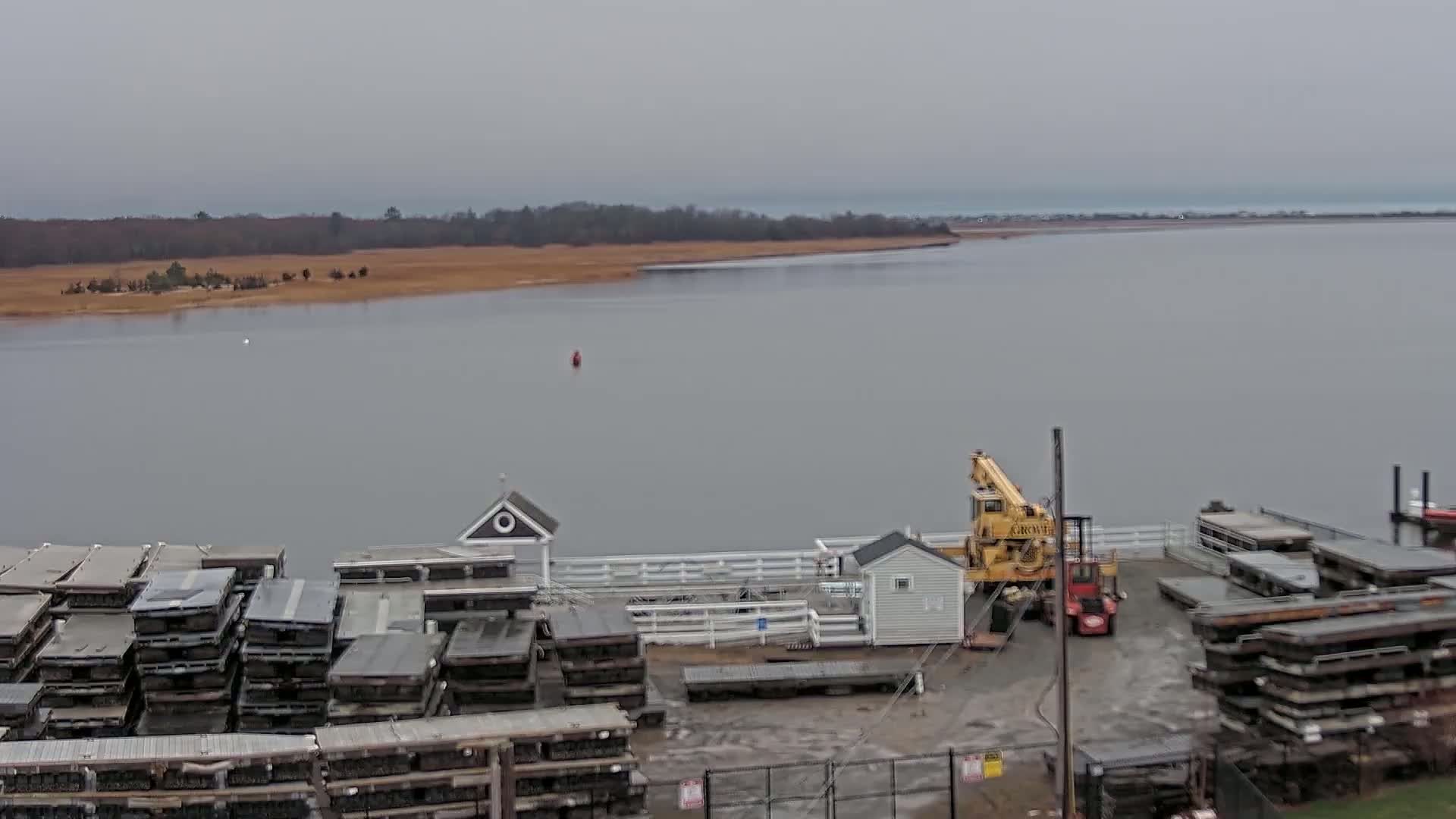 A gray, overcast day reveals a calm body of water bordered by land with low-lying vegetation, a small dock with stacked floating docks, a crane, and small buildings.