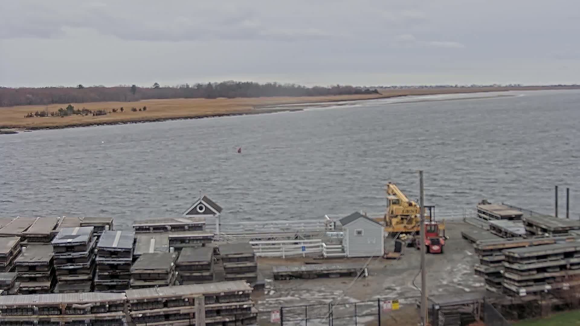 A choppy body of water stretches out to a distant shoreline under an overcast sky, with a dock holding stacked materials and a small crane nearby.