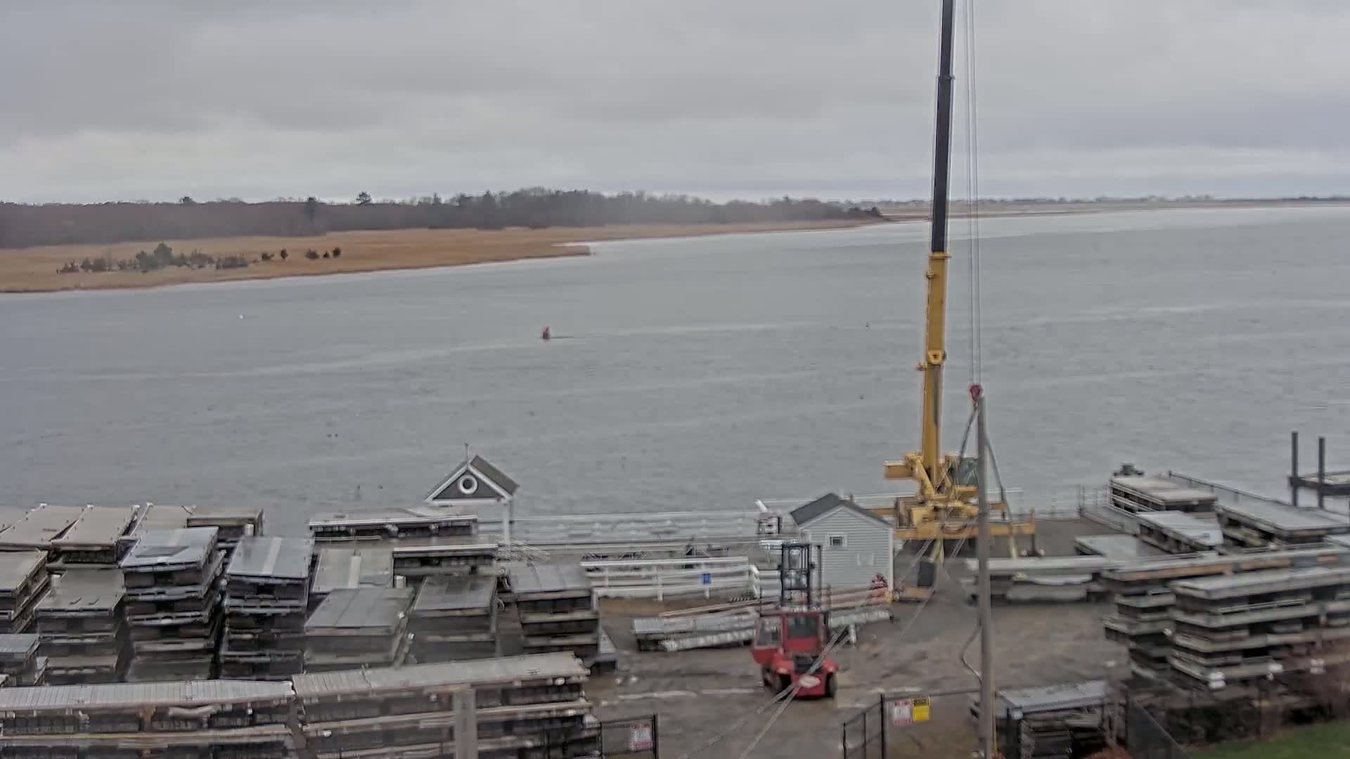 A yellow crane operates near stacks of gray metal components by a body of water under an overcast sky.