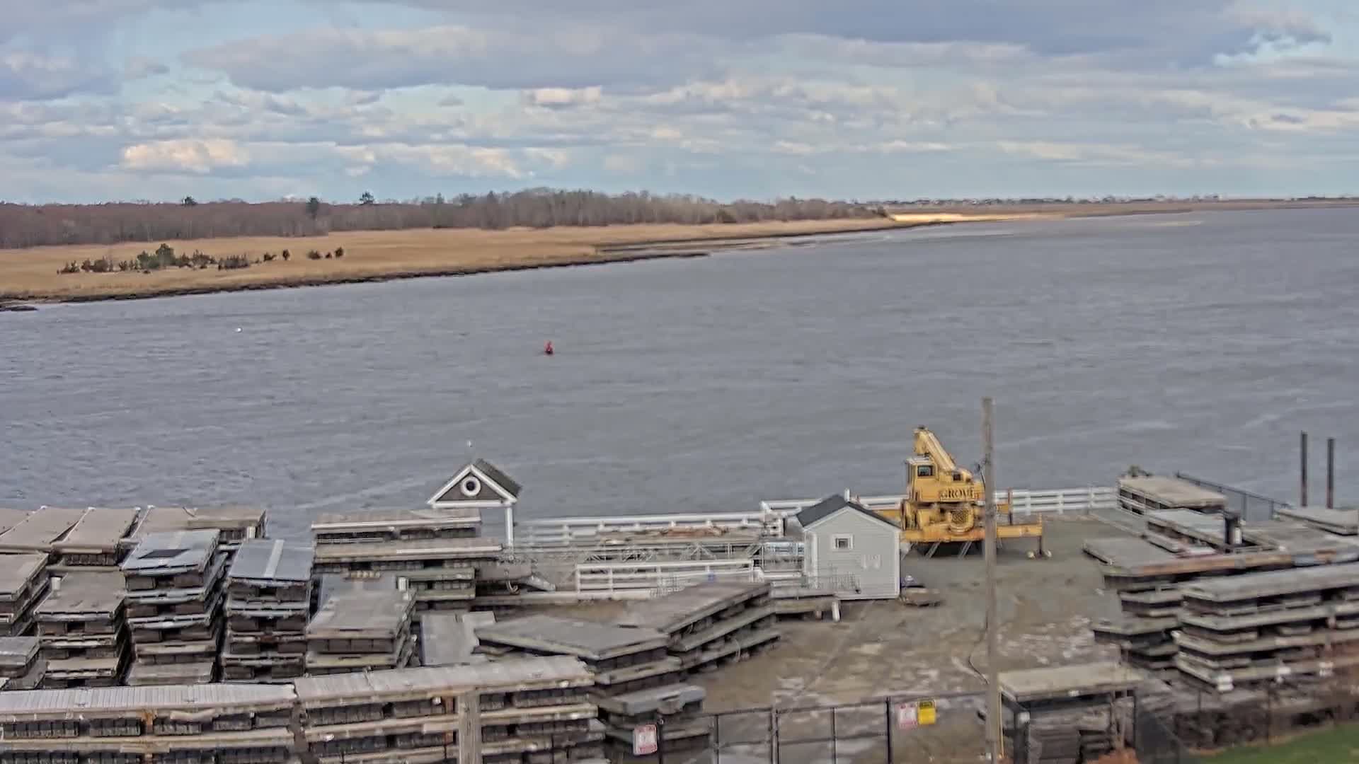 A yellow crane sits near stacks of floating docks by a calm body of water under a cloudy sky, with a distant shoreline of brown marsh grasses.