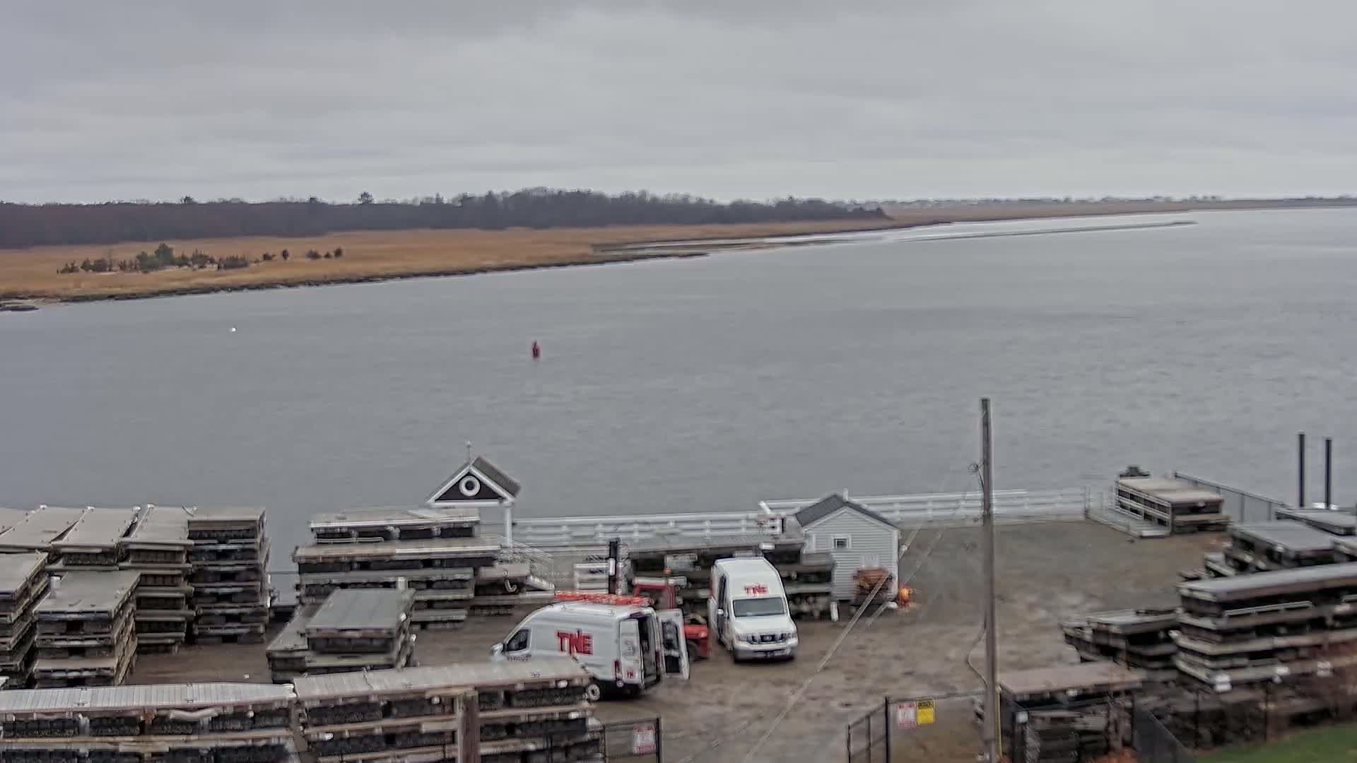 A calm, gray day overlooks a body of water with stacked dock parts and two white work vans parked near small buildings along the shore.