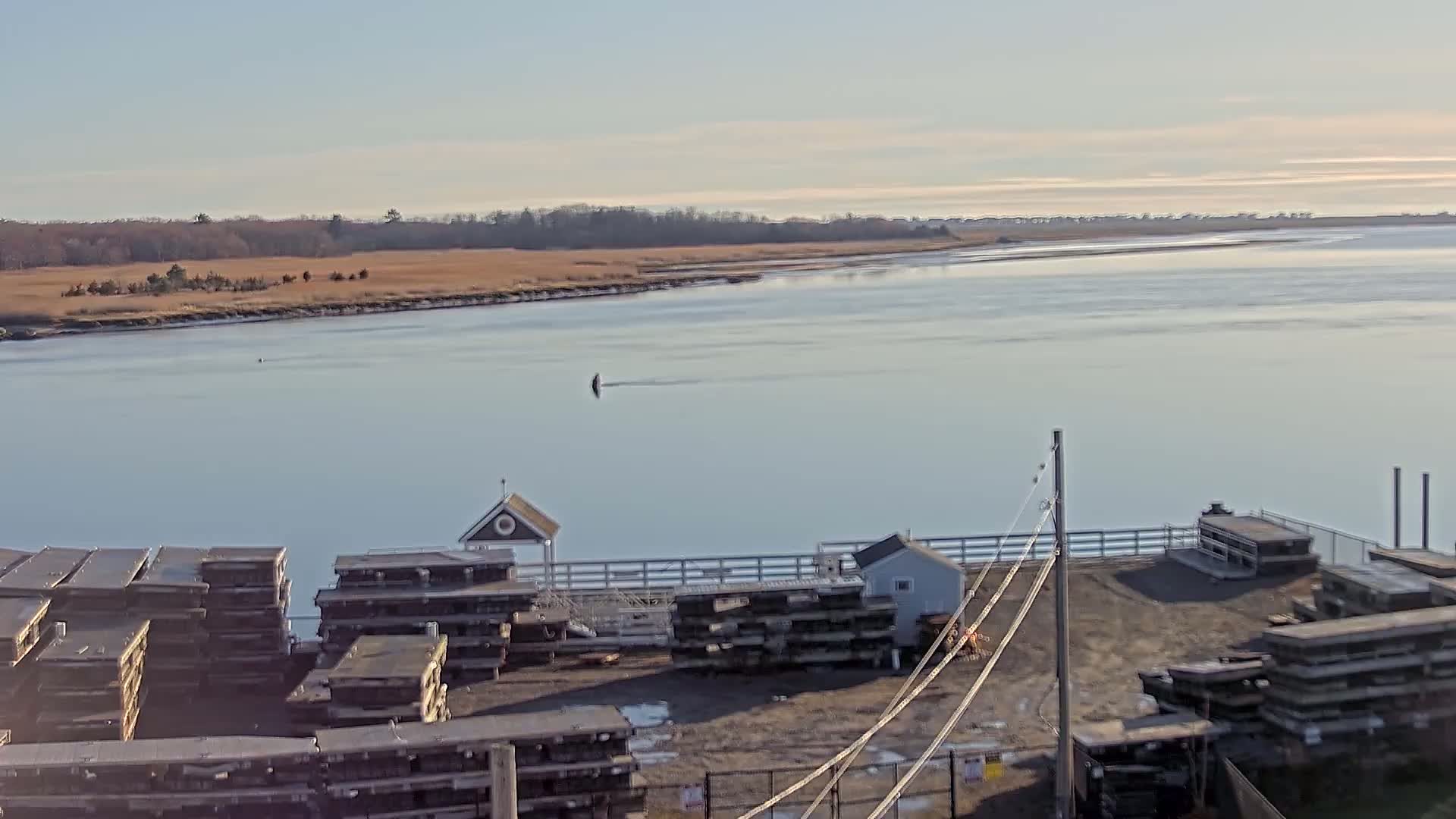 A calm, clear day reveals a wide body of water with a small structure and stacks of dark-colored materials near a shoreline of muted brown grasses and distant trees.